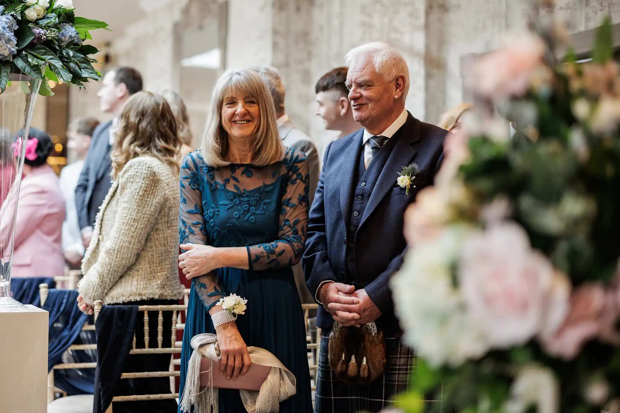 An older woman in a blue lace dress and an older man in a kilt and blazer stand smiling among seated guests at a wedding ceremony, surrounded by floral arrangements and soft lighting, with blurred people and flowers in the background.