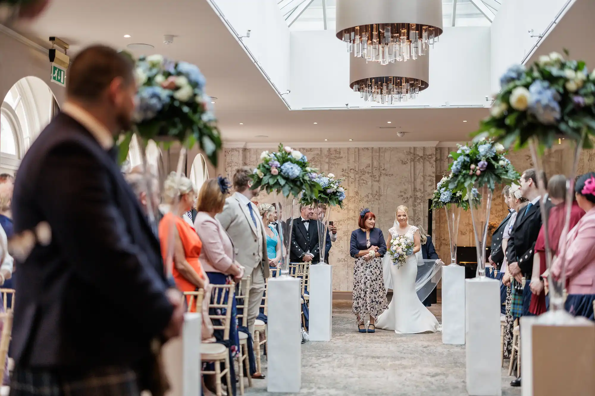 A bride in a white dress, holding a bouquet, walks down the aisle with an older woman. Guests in formal attire sit and stand on both sides, surrounded by tall floral arrangements. The setting is elegant with neutral tones and a modern chandelier overhead.