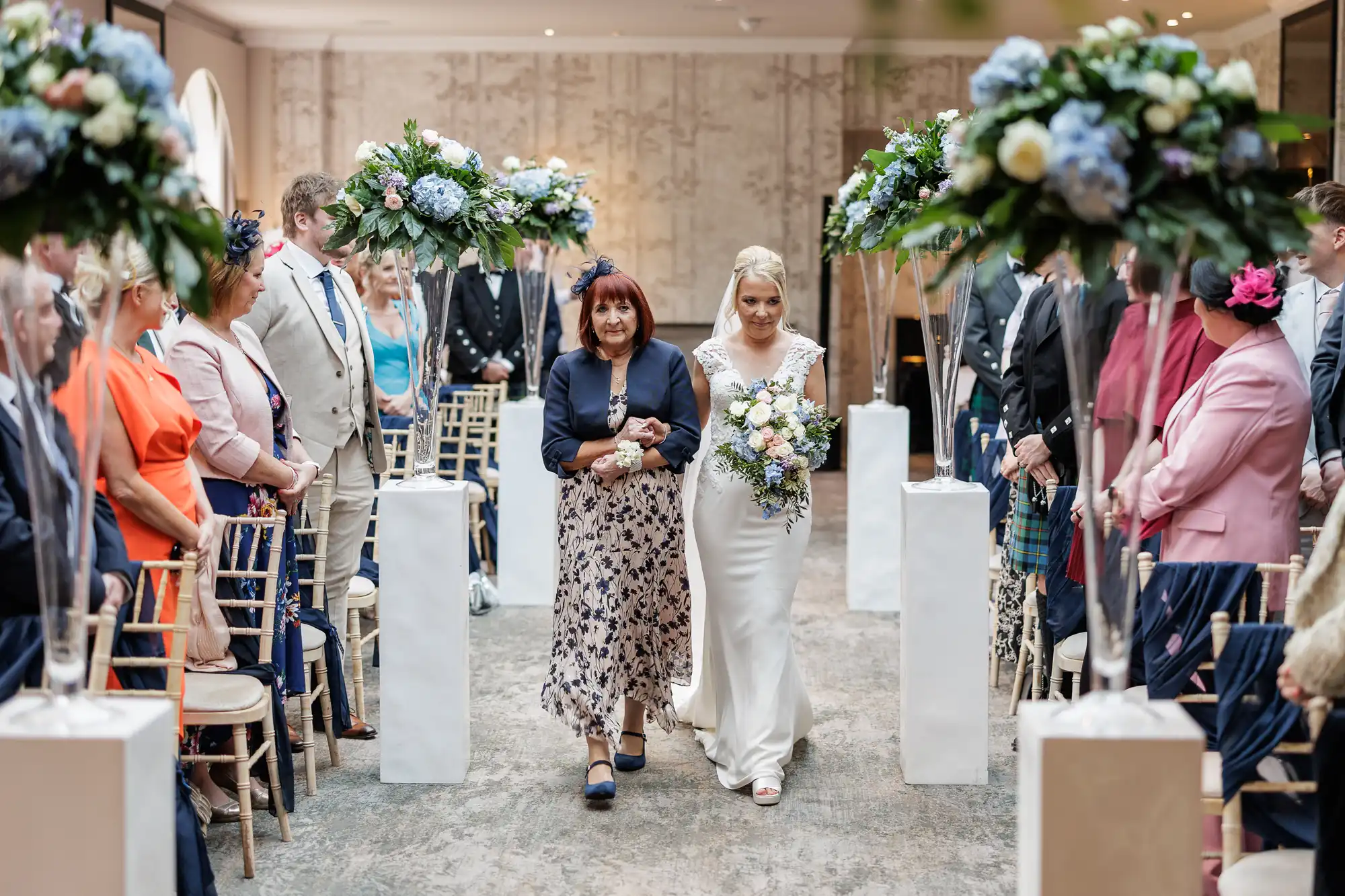 A bride in a white dress holding a bouquet walks down the aisle with an older woman. Guests sit on either side, some in colourful outfits. Tall floral arrangements line the aisle in a bright, elegant venue.