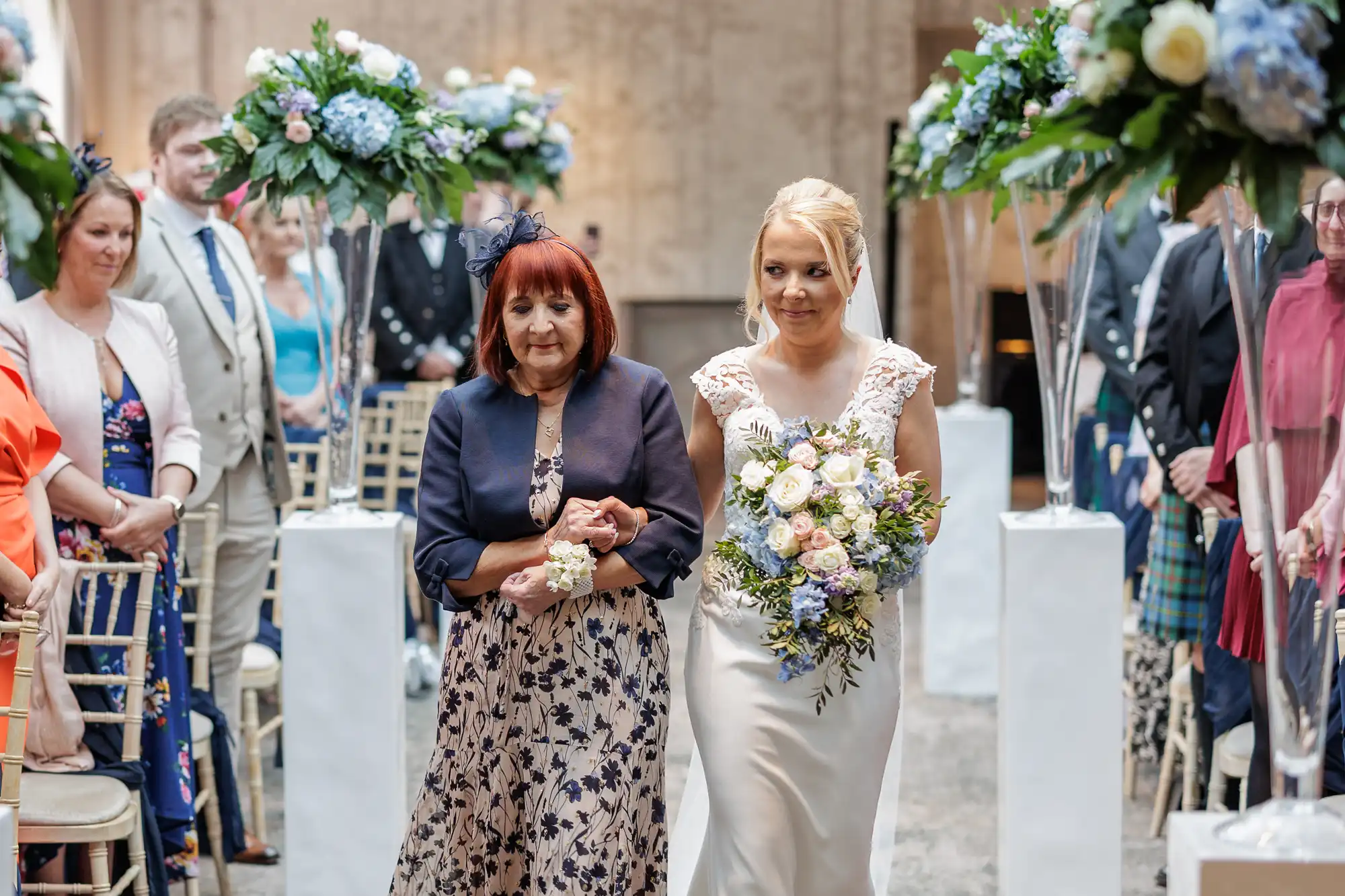 A bride in a white gown holding a bouquet walks down an aisle with an older woman in a navy jacket and floral dress. Guests seated on gold chairs watch, surrounded by tall floral arrangements of blue and white flowers on white pillars.