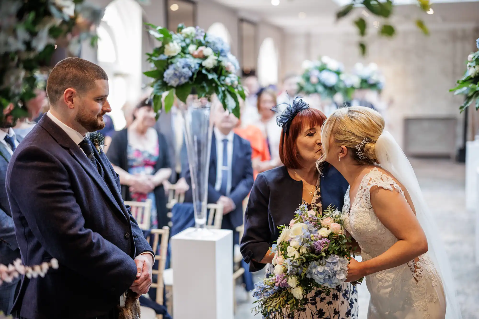 A bride in a white gown and veil kisses an older woman with red hair during a wedding ceremony. The groom, in a dark suit, stands nearby smiling. Guests and tall floral arrangements with blue and white flowers are visible in the bright, elegant room.