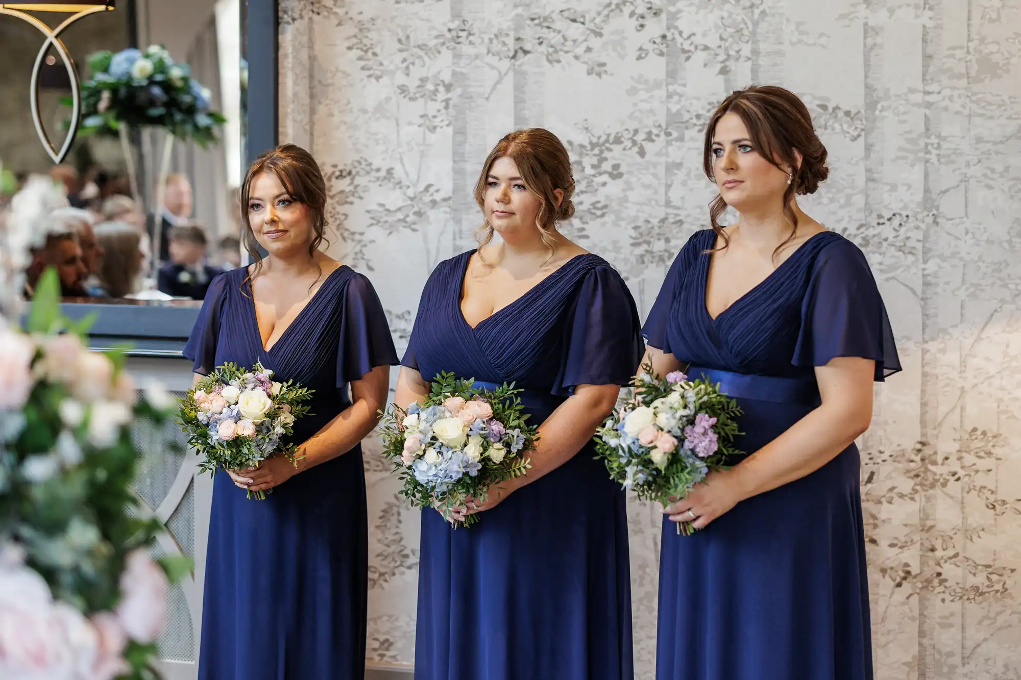 Three women in matching navy blue dresses stand indoors, holding bouquets of pink and white flowers with greenery. They face left, appearing attentive. The background features a light, patterned wall and a mirror reflecting people seated at the event.