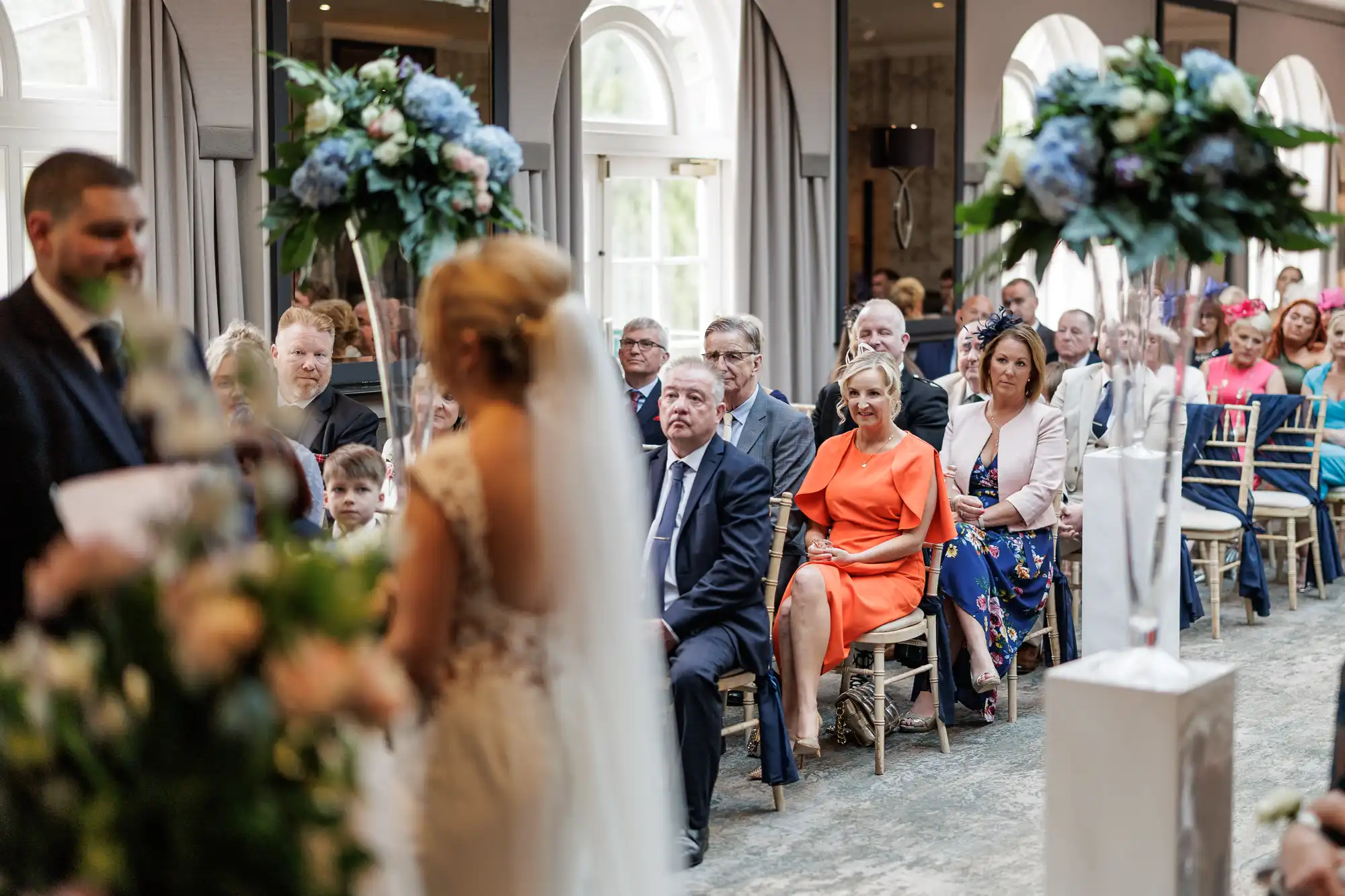 A bride in a white dress faces a man, possibly the officiant, during an indoor wedding ceremony. Guests sit in rows of chairs, watching attentively. Tall floral arrangements with blue and white flowers decorate the aisle. Large arched windows let in natural light.
