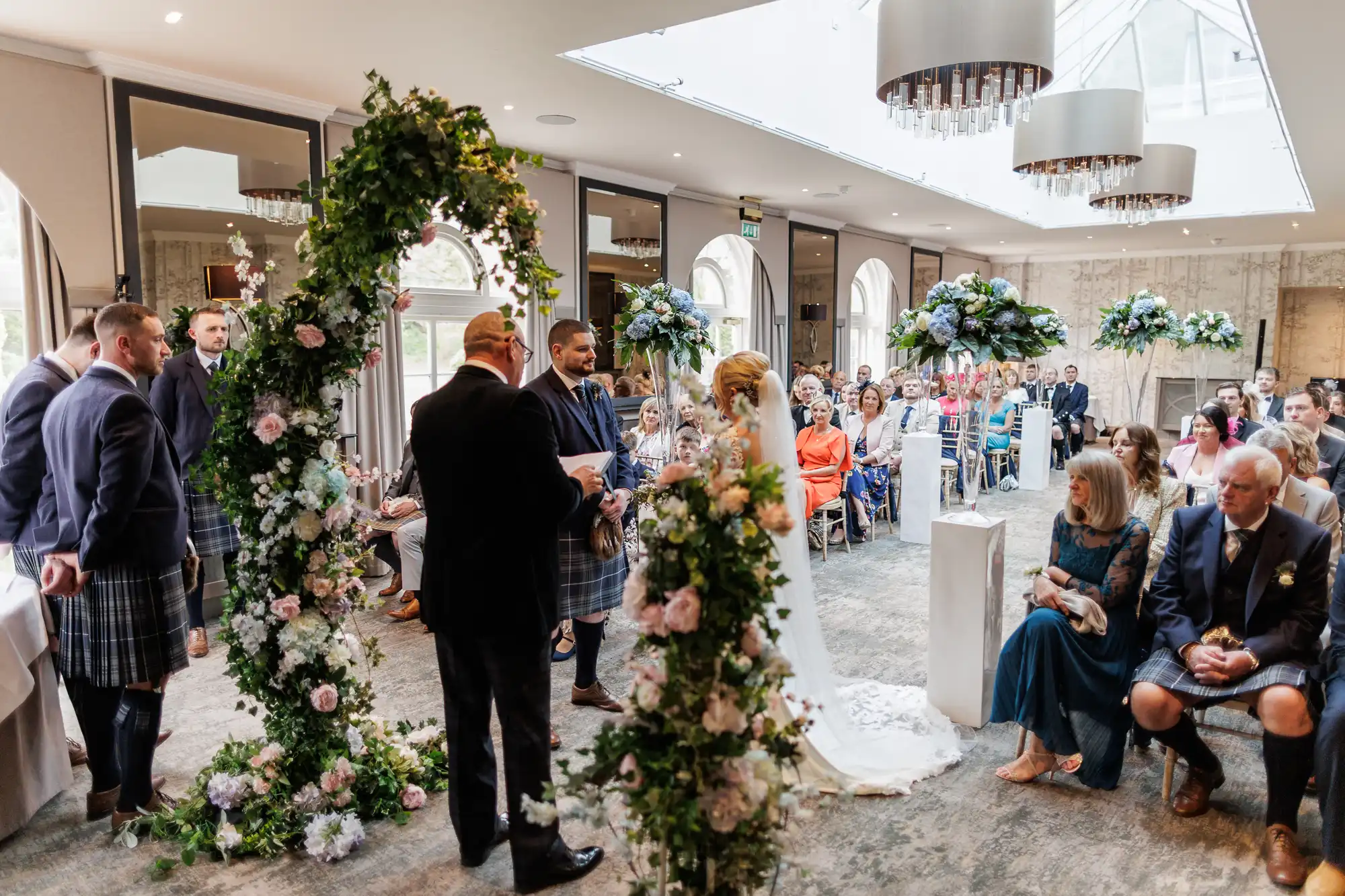 An indoor wedding ceremony with guests seated and watching a couple stand under a flower-covered arch. The groom and groomsmen wear kilts, while the bride wears a white gown and veil. The room has elegant lighting and large floral arrangements on tables.