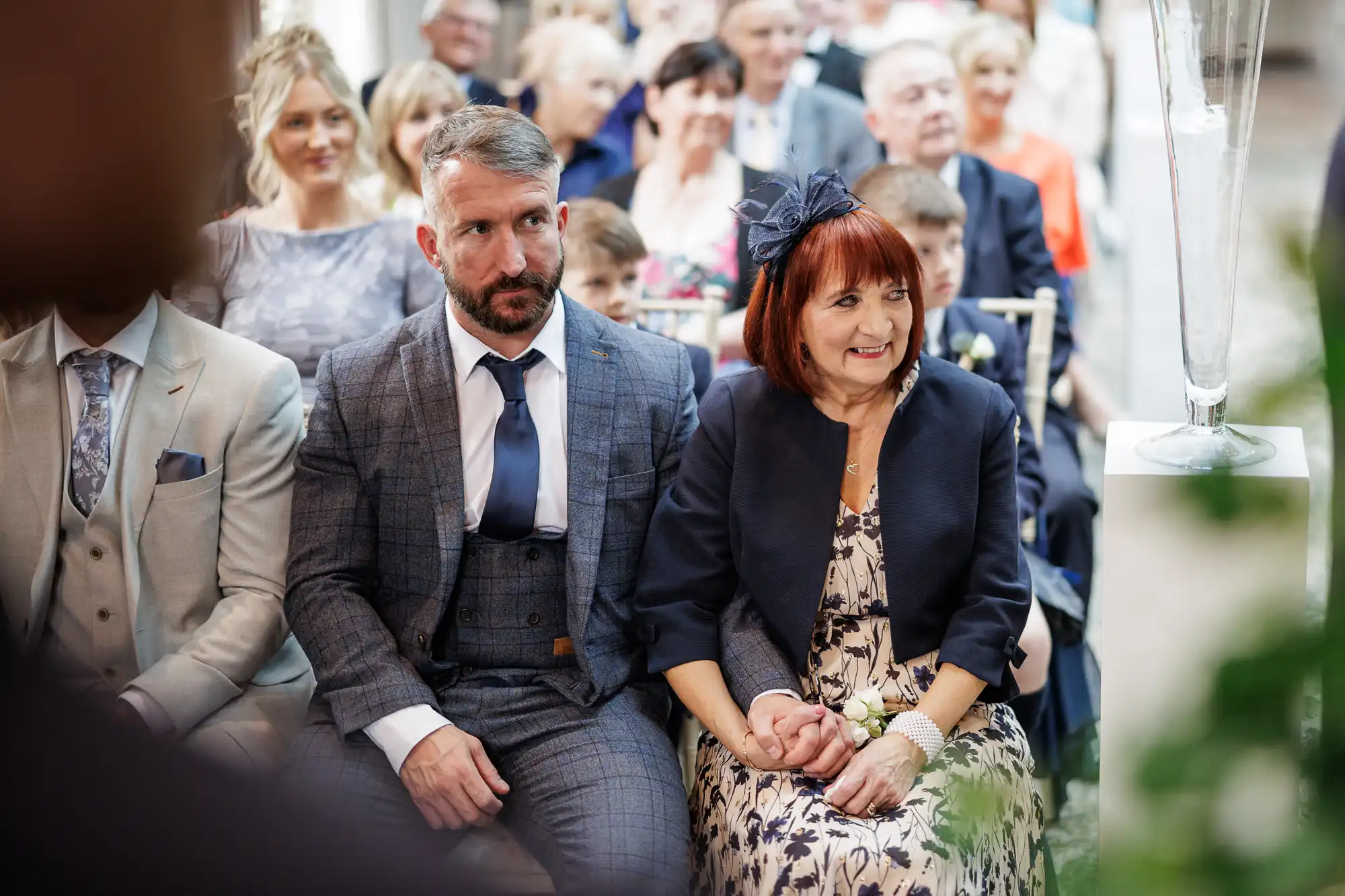 A man in a grey suit and a woman in a navy floral dress sit holding hands at a wedding ceremony, looking ahead. Other well-dressed guests, including children, are seated behind them, all focused on the event. The setting is bright and formal.