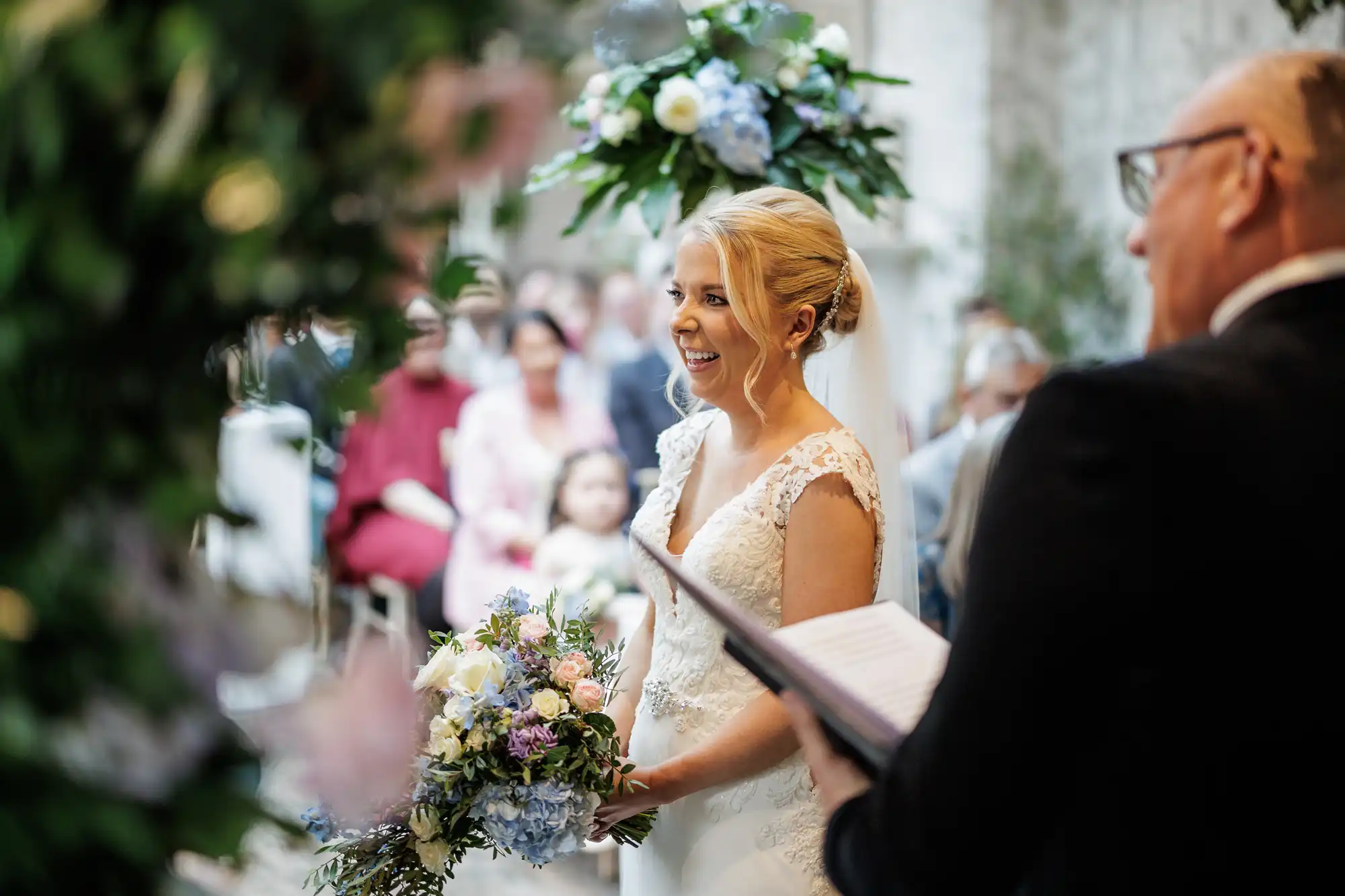 A smiling bride in a lace white gown holds a pastel bouquet during a wedding ceremony, standing beside an officiant with an open book. Guests are seated in the background, and floral arrangements decorate the scene. The atmosphere is joyful and elegant.