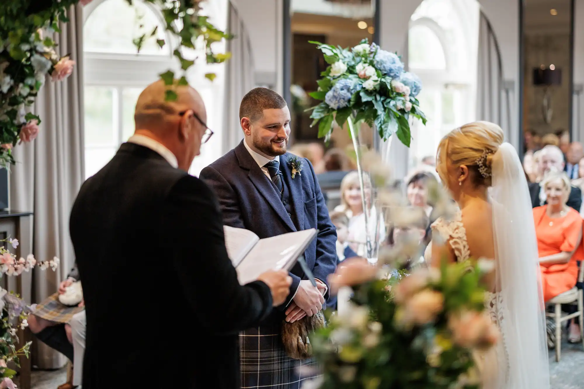 A bride and groom stand facing each other during an indoor wedding ceremony. The officiant reads from a book. The groom smiles warmly, and guests are seated in the background among floral decorations and natural light.