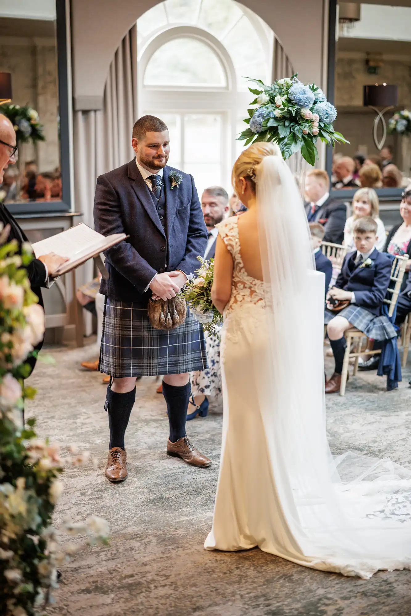 A bride in a white dress with a long veil stands facing a groom in a traditional Scottish kilt and dark jacket during an indoor wedding ceremony. Guests, some in kilts, are seated and watching. There are flower arrangements and an officiant holding a book.