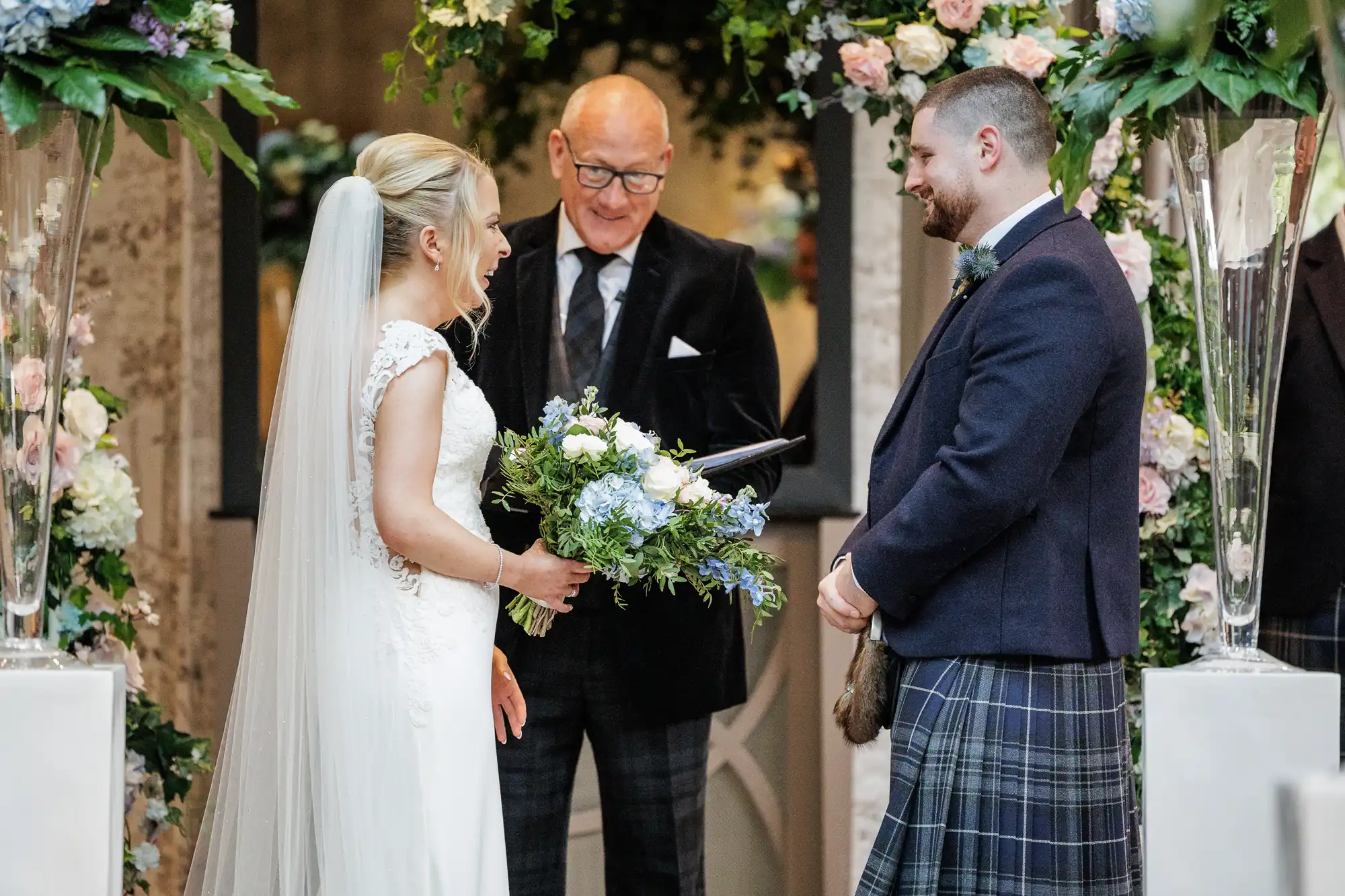 A bride in a white dress holds a bouquet of blue and white flowers, facing a smiling groom in a dark jacket and tartan kilt. An officiant stands between them, and they are surrounded by lush floral arrangements in soft pastel colours.