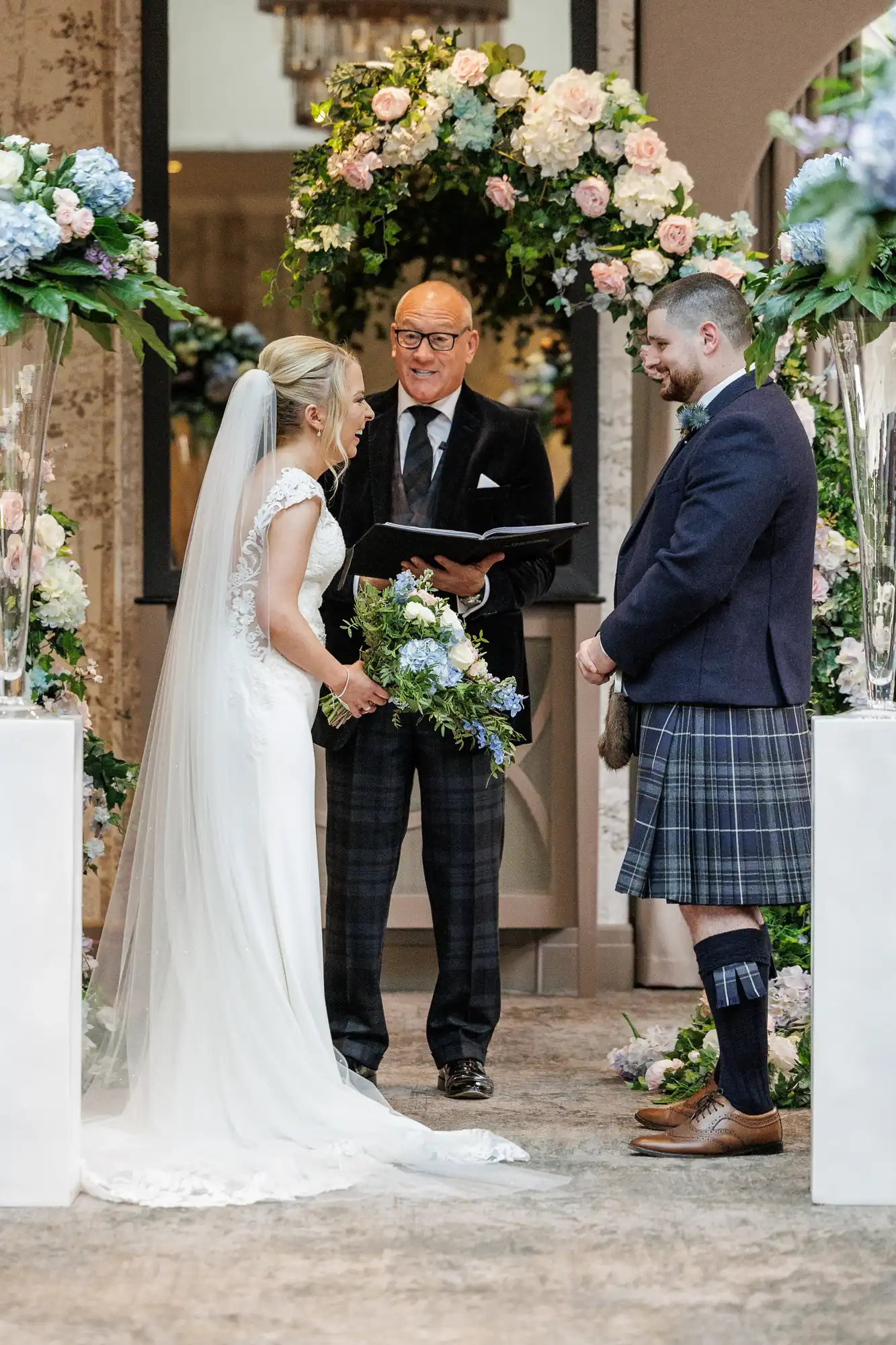 A bride in a white dress and veil holds a bouquet and faces a groom in a dark jacket and tartan kilt. They stand under a floral arch, smiling at an officiant holding a book, during an indoor wedding ceremony with lush flower arrangements.