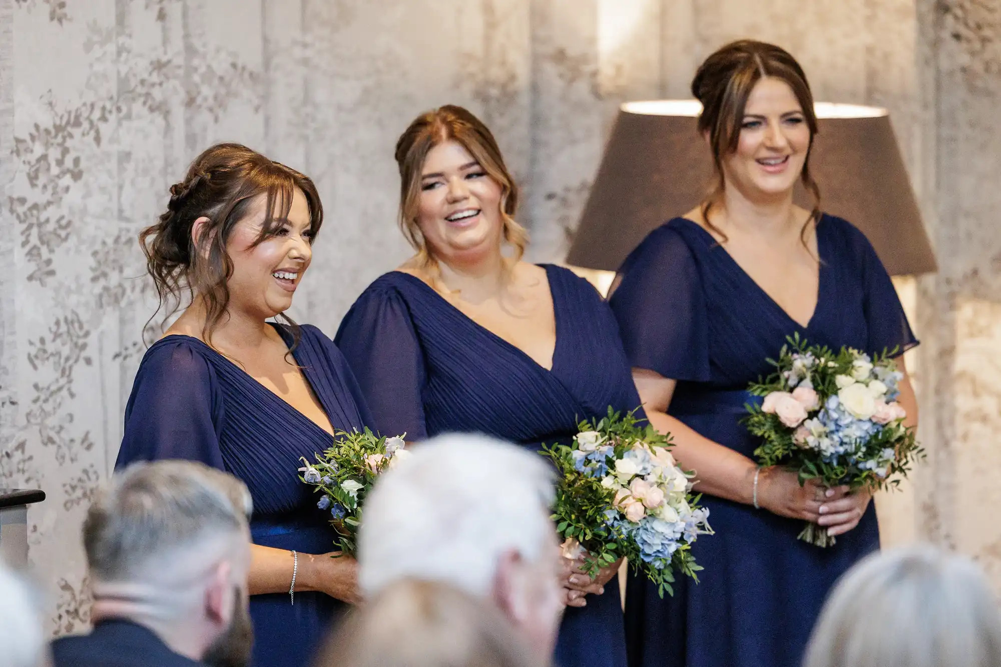 Three bridesmaids in matching navy blue dresses stand indoors, smiling and holding floral bouquets with greenery and white flowers. They face a seated audience, with a softly-lit lamp and patterned wallpaper in the background.