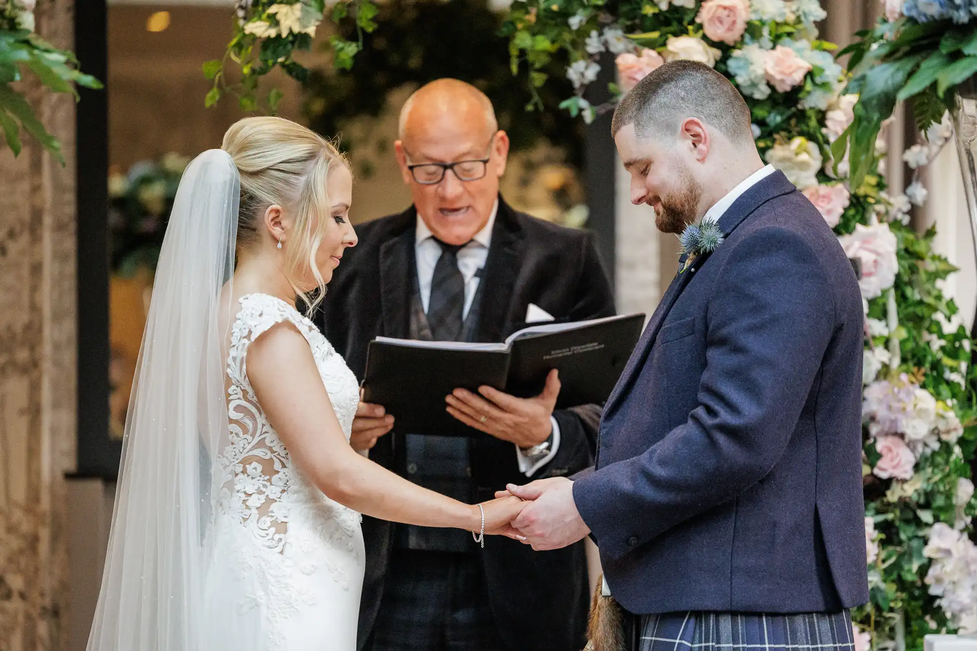 A bride and groom stand facing each other, holding hands, during a wedding ceremony. The officiant reads from a book behind them. The bride wears a white dress with lace details and a veil; the groom wears a dark jacket and kilt. Floral arrangements decorate the background.