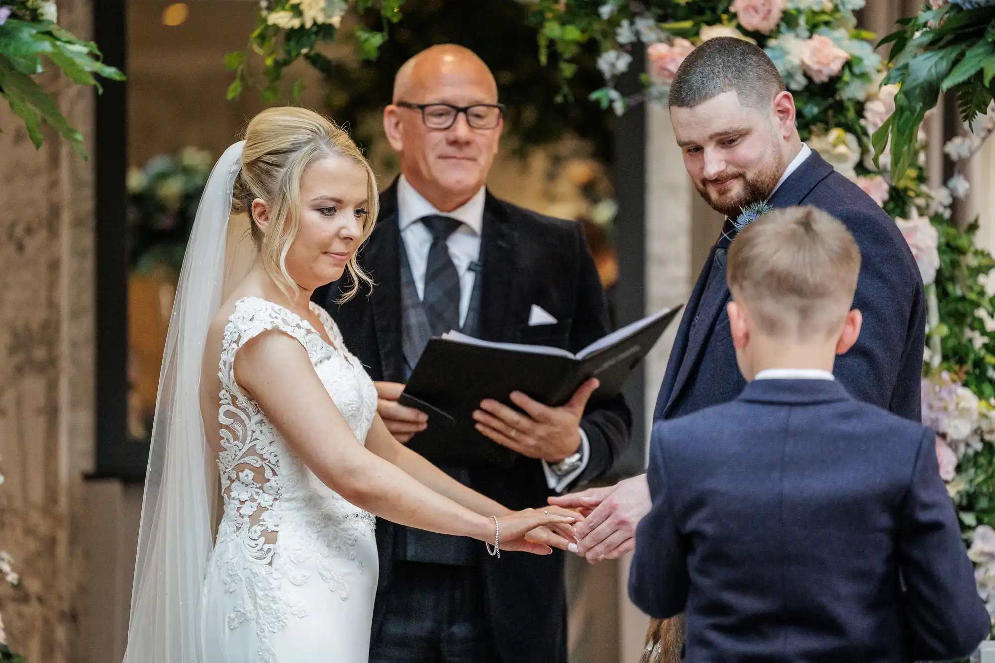 A bride in a white lace gown and veil holds hands with a groom in a dark suit as an officiant reads from a book behind them. A young boy stands in front, participating in the ceremony. Floral decorations and greenery surround the group.