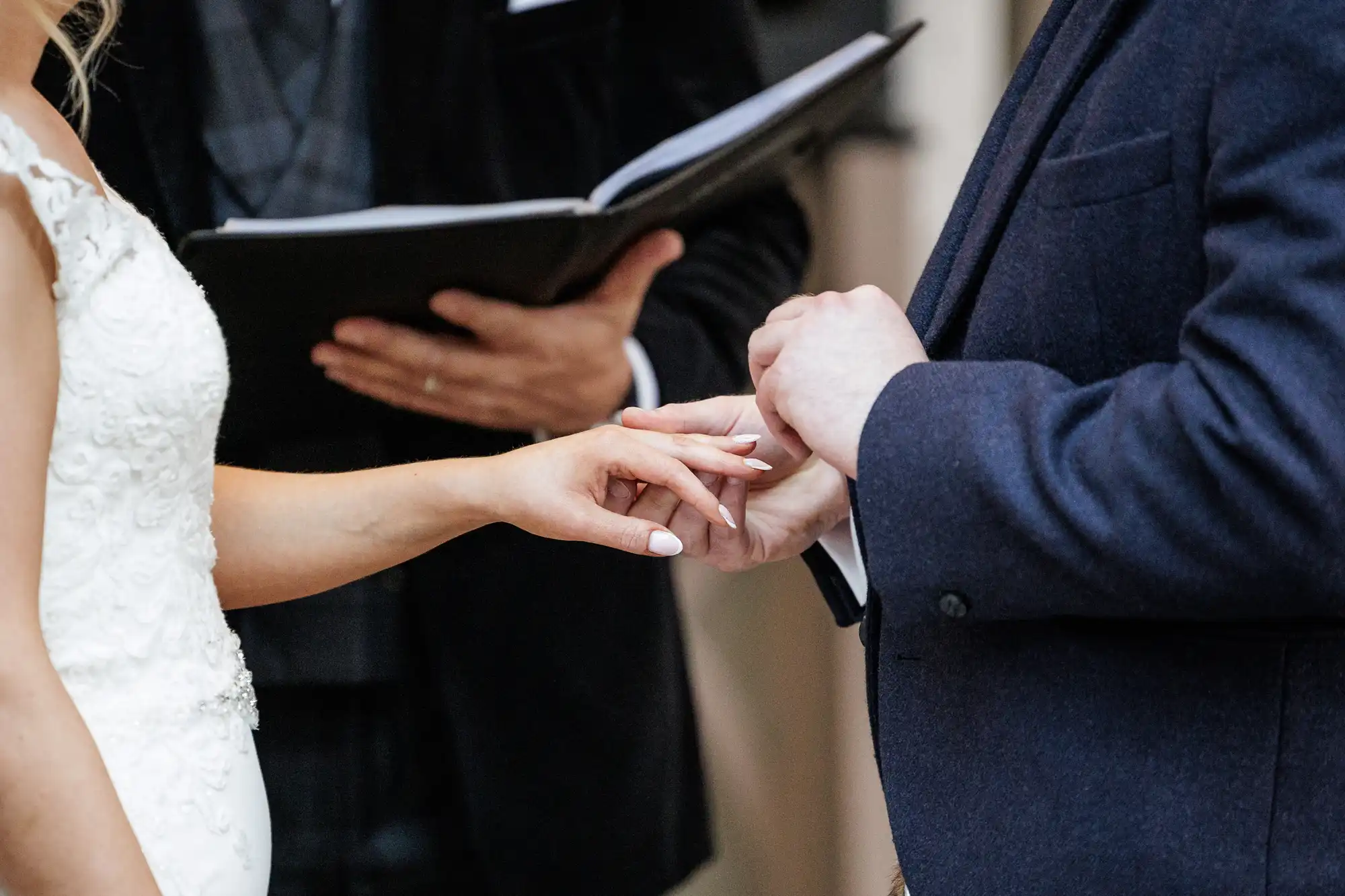 A close-up of a bride placing a wedding ring on the groom’s finger during a ceremony. The bride wears a white dress, and the groom wears a dark suit. An officiant holding an open book stands in the blurred background.