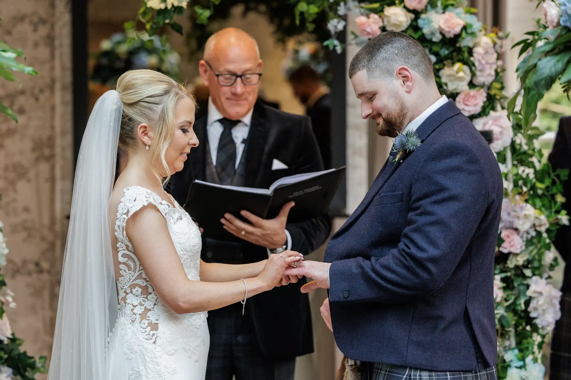 A bride in a white lace gown and veil places a ring on the groom’s finger during their wedding ceremony. The groom wears a dark suit and tartan kilt. An officiant in spectacles reads from a book behind them, with floral arches framing the scene.