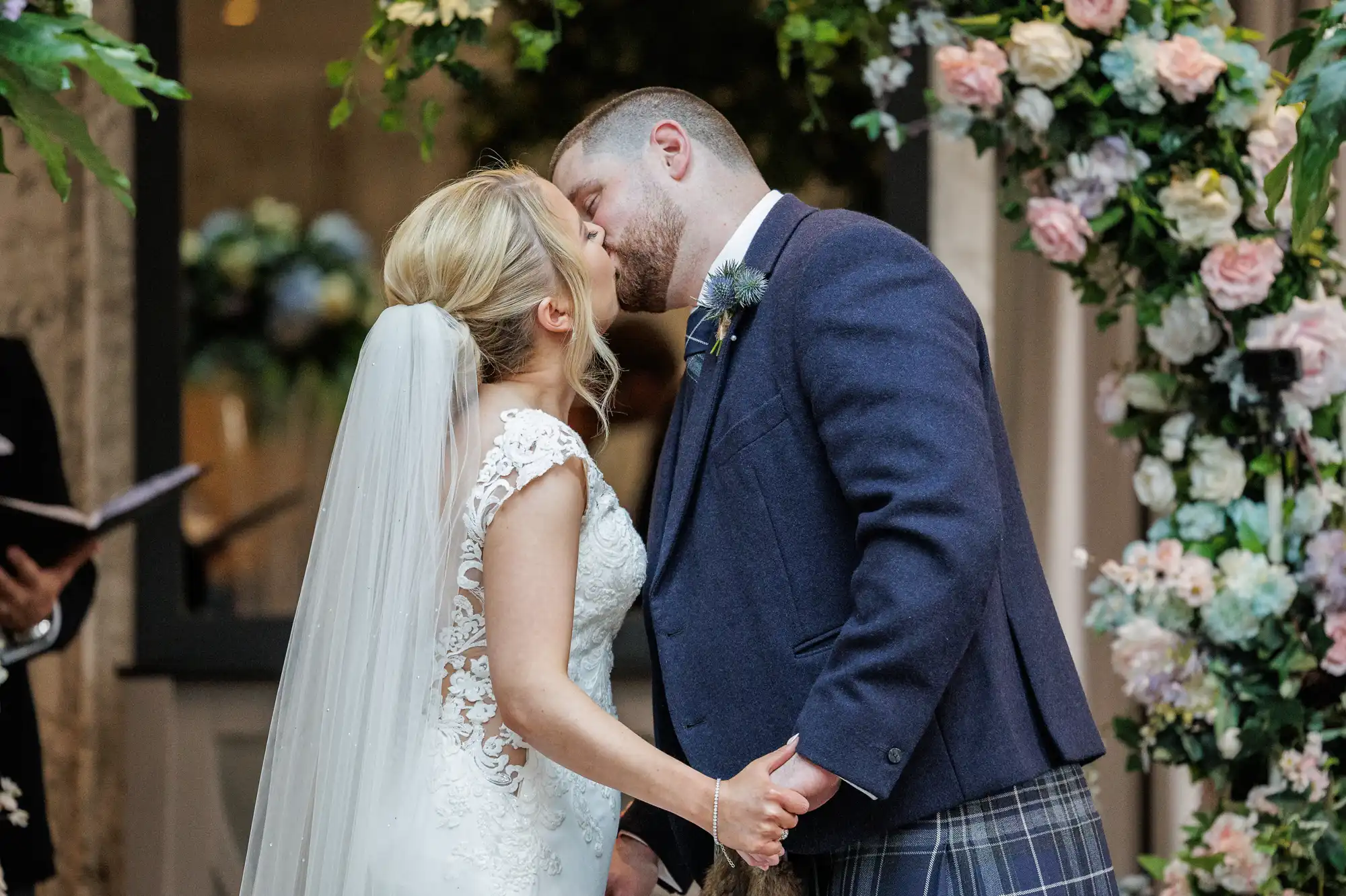 A bride and groom share a kiss during their wedding ceremony. The bride wears a lace gown and veil, whilst the groom wears a navy jacket and tartan kilt. They hold hands beneath a flower arch decorated with pink and white blooms.