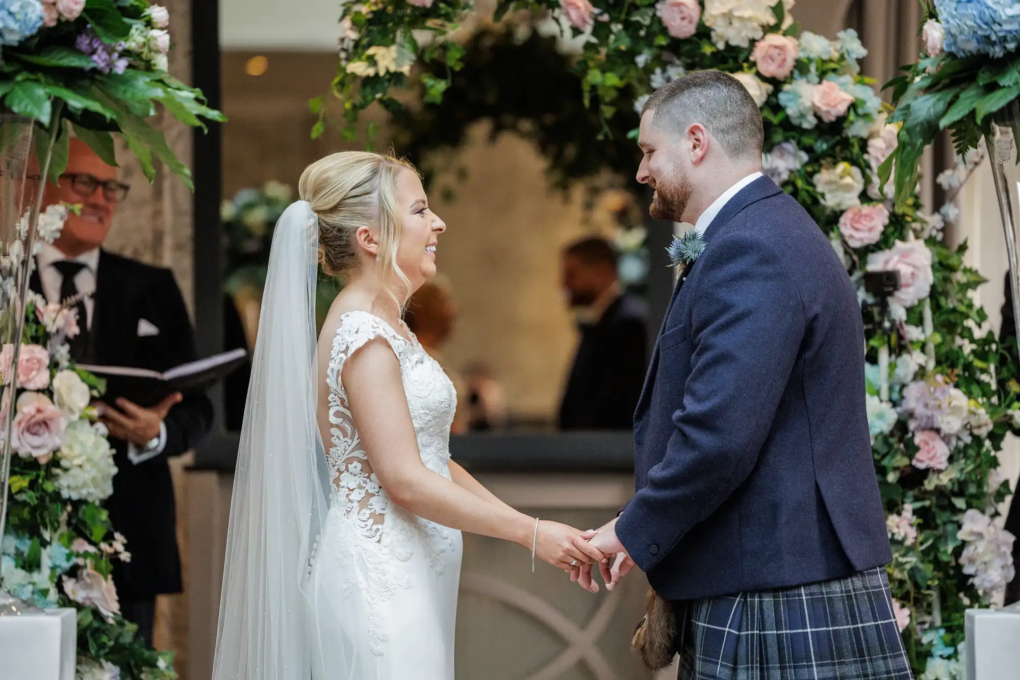 A bride in a white dress and veil smiles as she holds hands with a groom in a dark jacket and checked kilt. They stand under a flower arch, facing each other. An officiant is in the background, holding a book. The scene appears joyful and romantic.