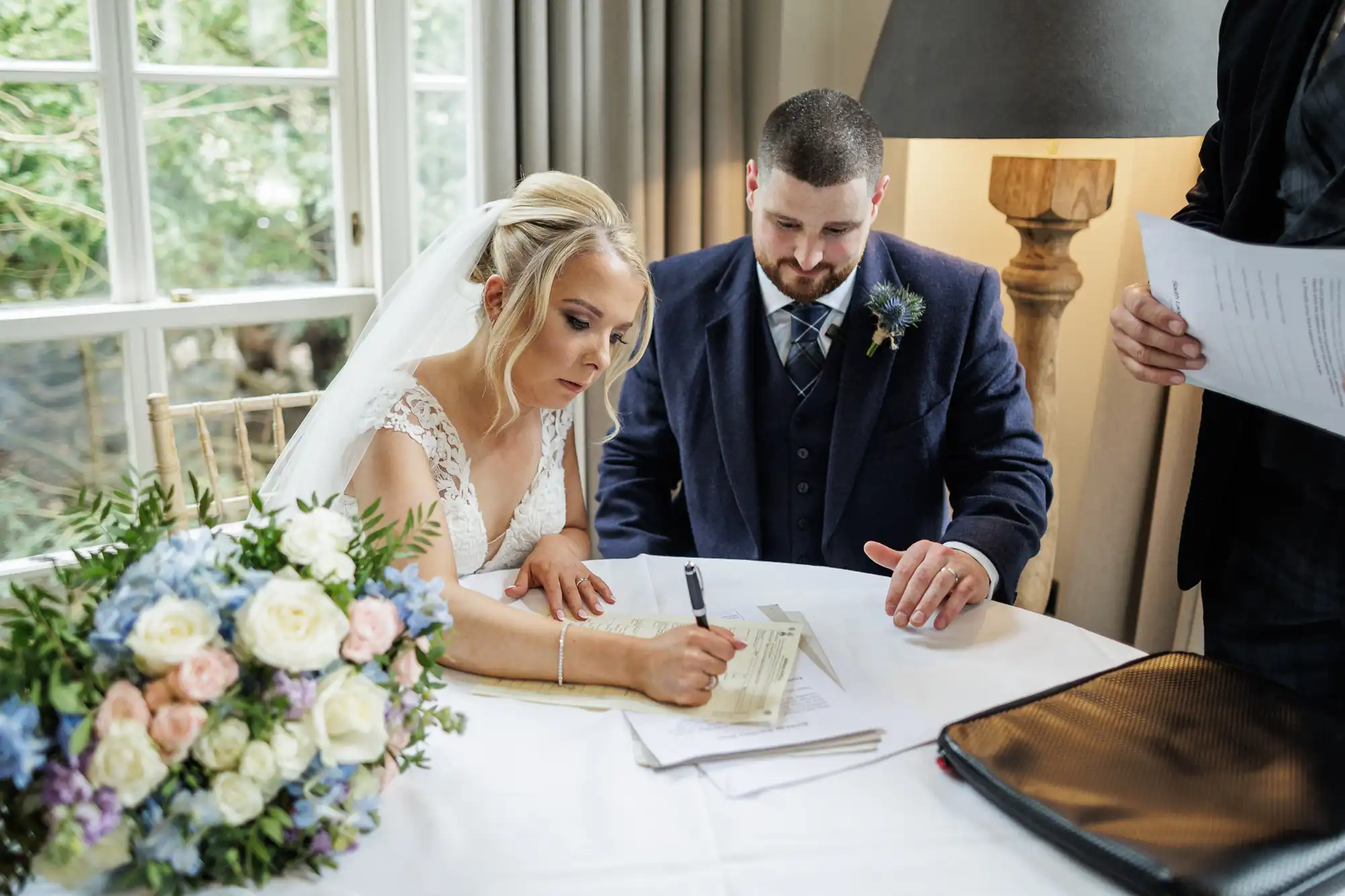 A bride in a white dress and veil signs a document at a table, while a groom in a navy suit watches. A bouquet of pastel flowers rests nearby. Another person holds papers beside them. Sunlight filters through a window and curtains in the background.