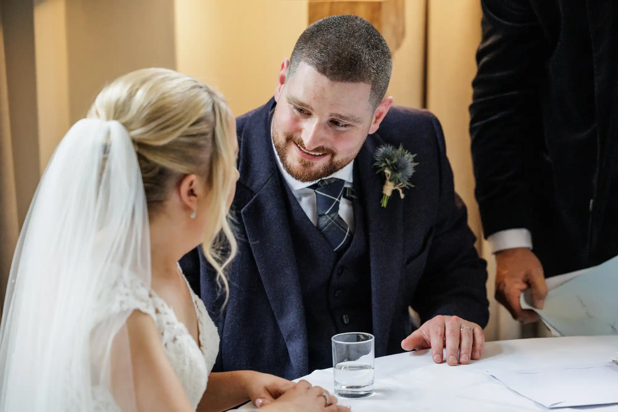 A bride in a white dress and veil sits at a table opposite a groom in a navy suit and checked tie. They smile warmly at each other. A glass of water sits on the table, and a person in dark clothing stands nearby, partially visible.
