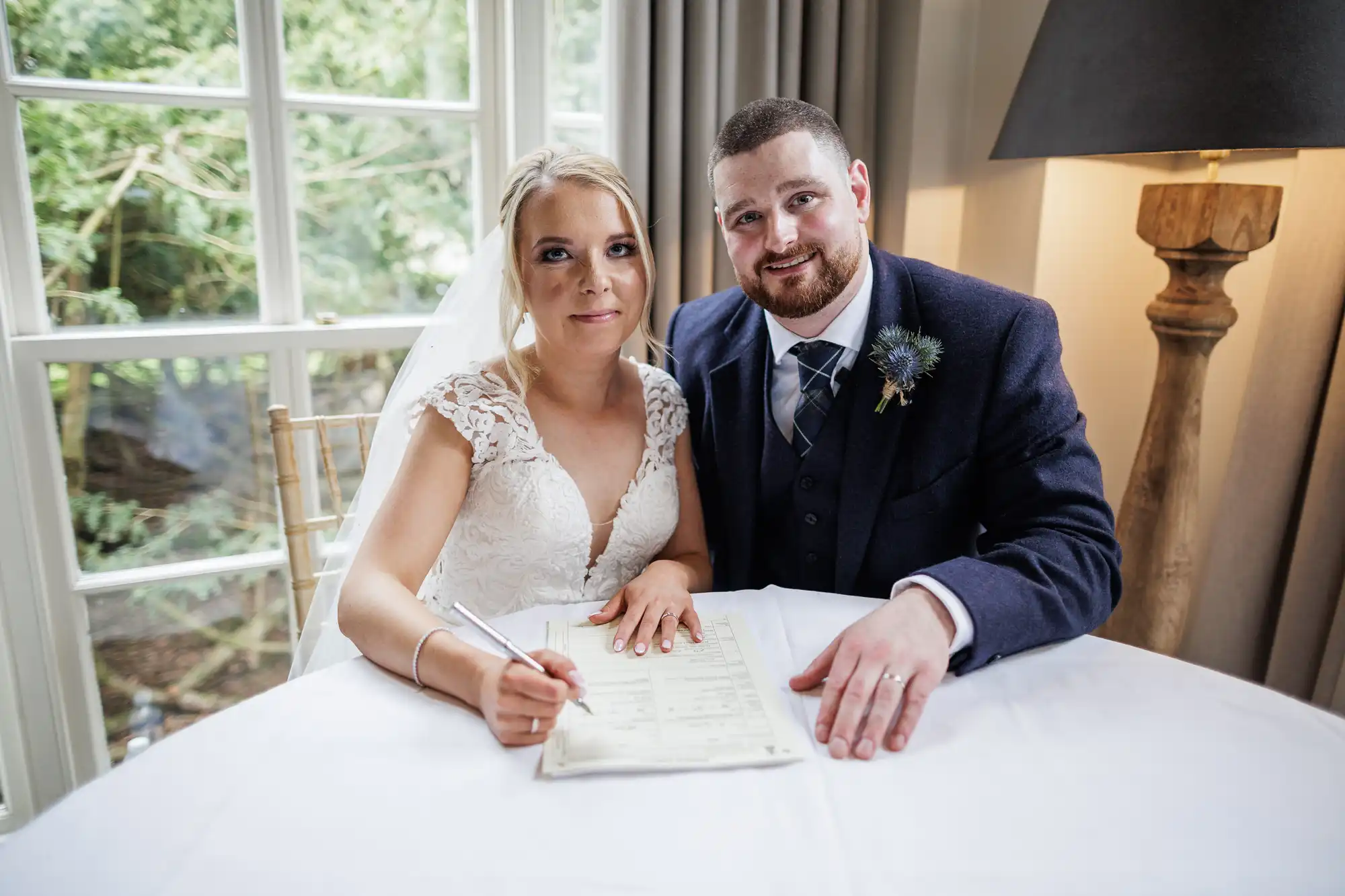 A bride in a white lace dress and veil sits beside a groom in a dark suit and checked tie. Both smile at the camera while she signs a document on a round table. They sit indoors by a window with greenery visible outside. A lamp and beige curtains are behind them.