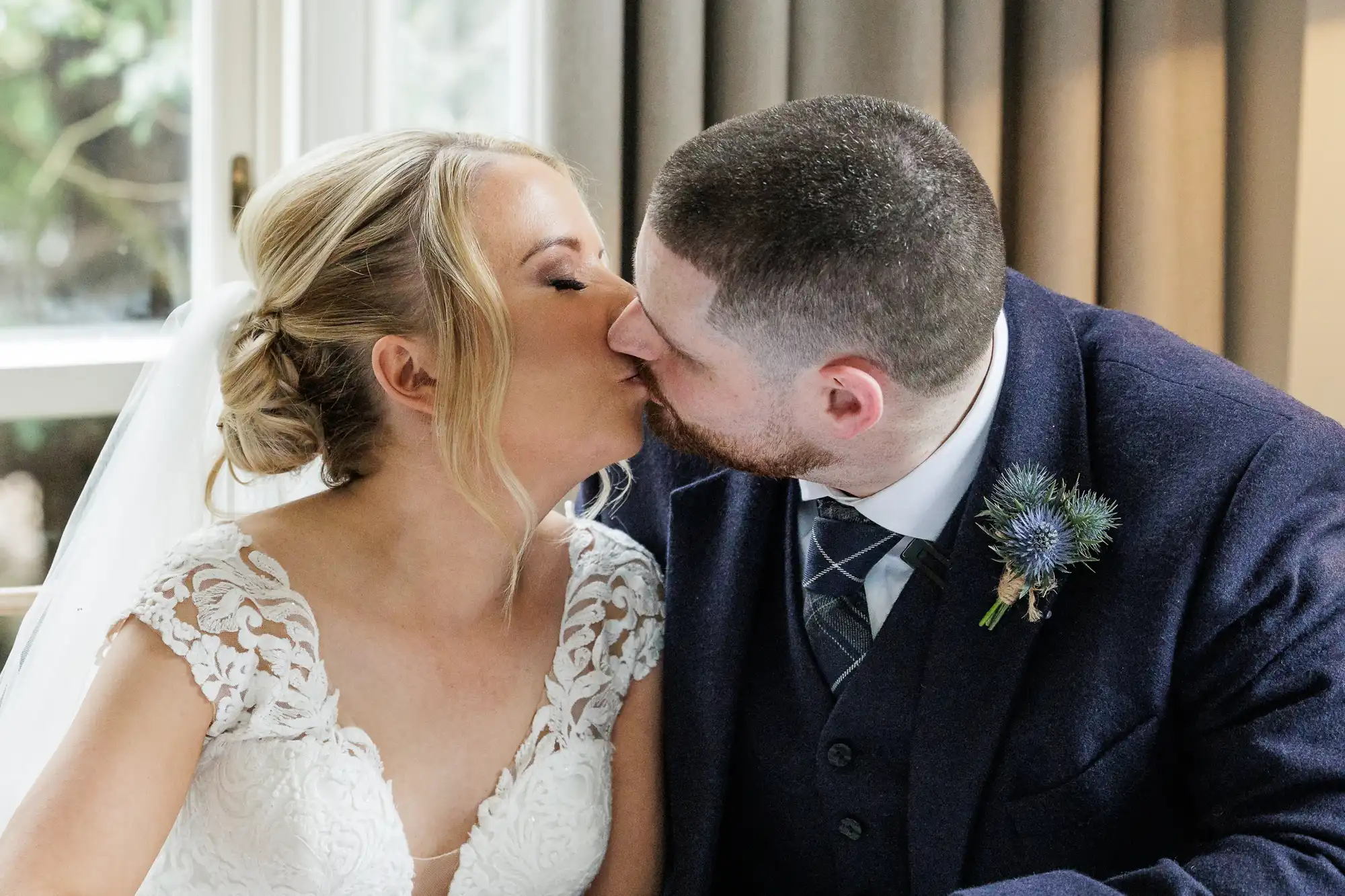 A bride in a white lace wedding dress and a groom in a navy suit with a thistle buttonhole share a kiss indoors, seated closely together. Light streams through a window behind them, and grey curtains are partially visible.