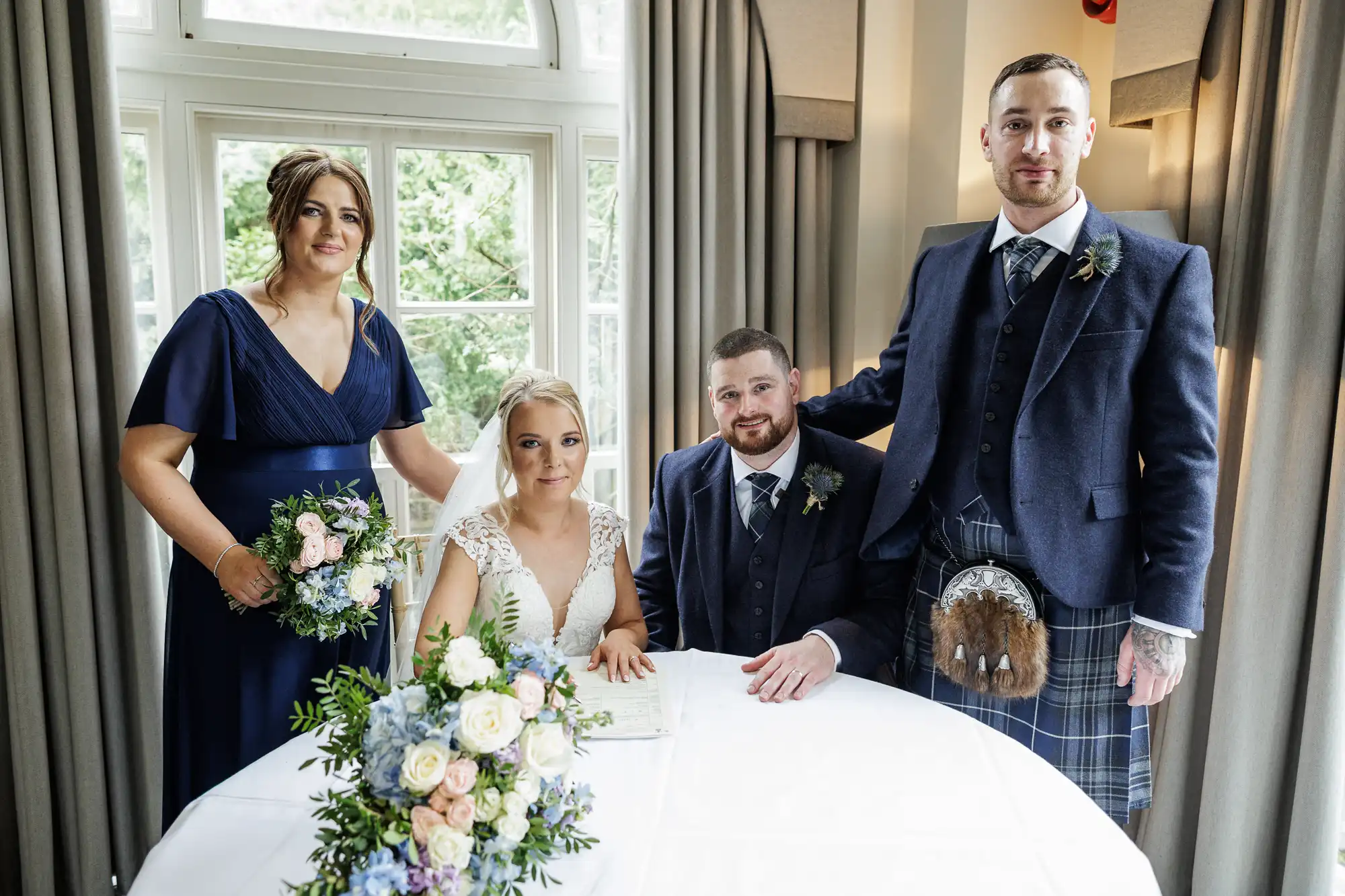 Four people pose at a wedding. A bride and groom sit at a table with a bouquet and flowers; the bride wears white, the groom a dark suit. Two attendants stand behind them: a woman in a navy dress holding flowers, and a man in a kilt and jacket. Large windows are behind them.