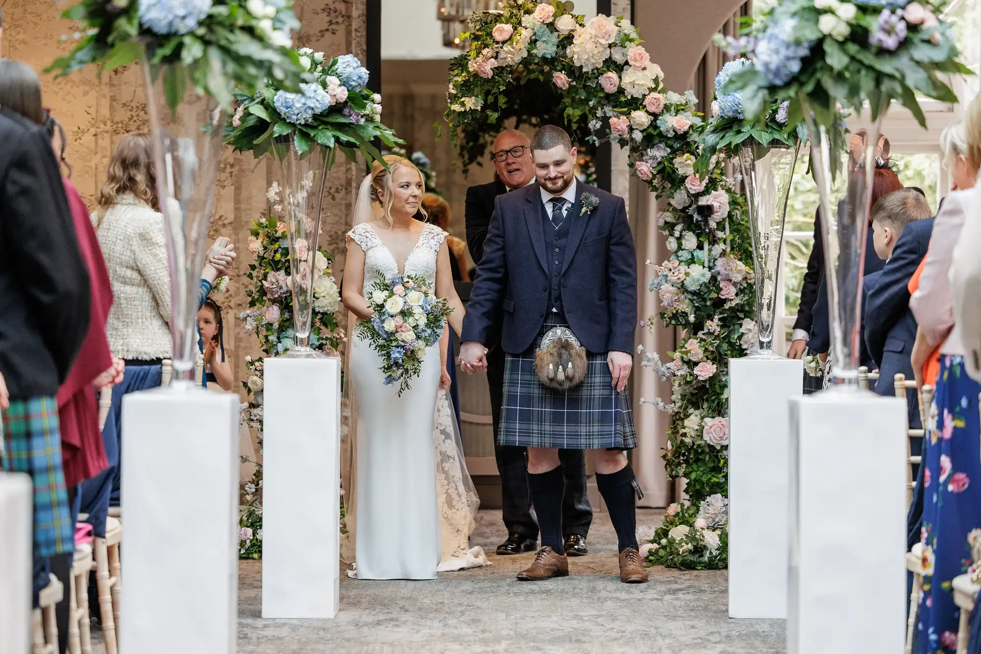 A bride in a white gown and a groom in a kilt stand together under a floral arch, holding hands and smiling, surrounded by guests seated on either side. The venue is decorated with white pillars and lush flower arrangements.