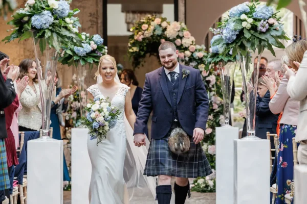 Balbirnie House wedding photos: A smiling bride in a white dress and a groom in a kilt and jacket walk hand-in-hand down an aisle lined with tall floral arrangements. Guests on both sides clap and cheer in a bright, flower-filled wedding venue.