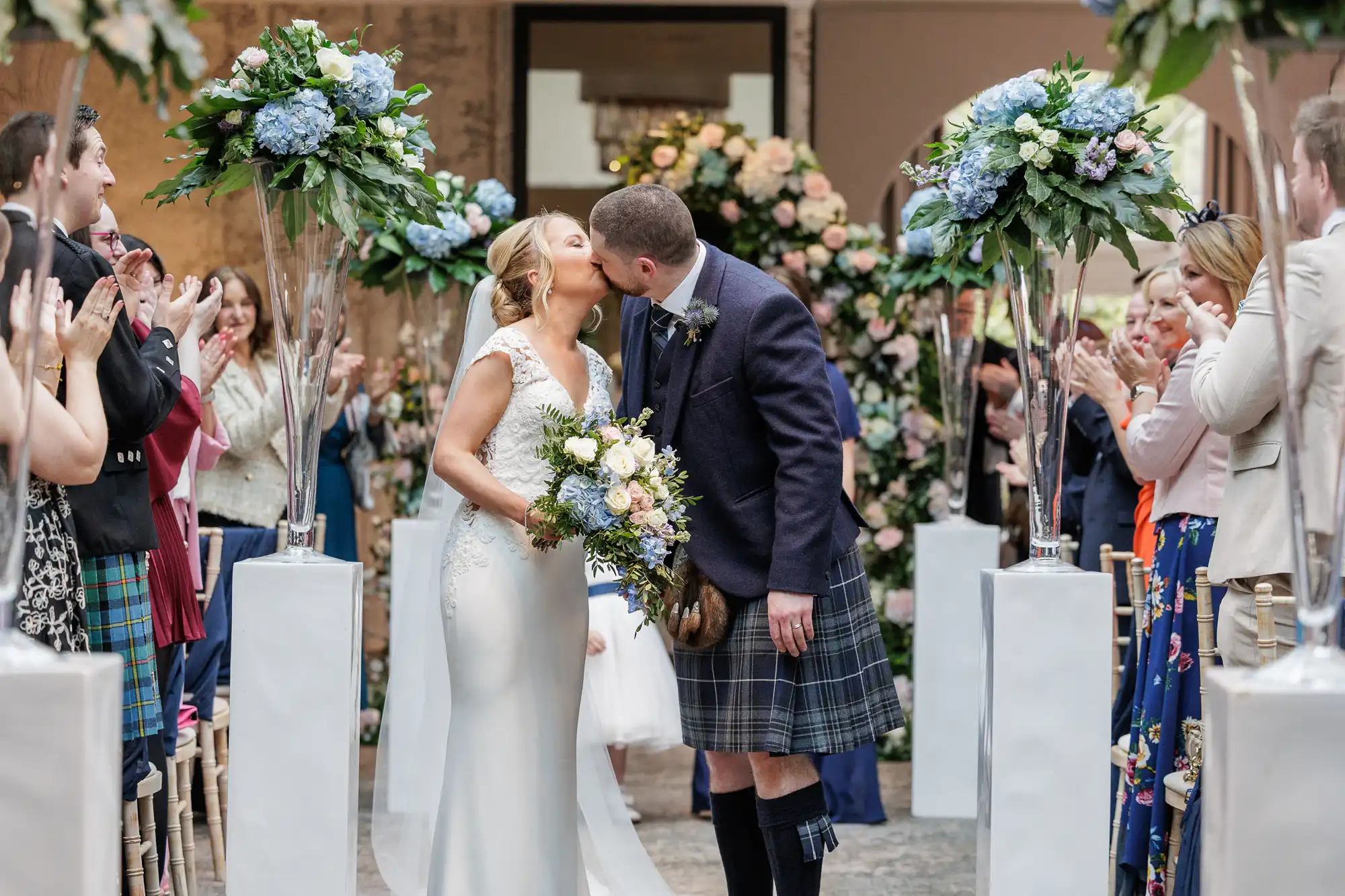 A bride and groom kiss at their wedding ceremony. The bride wears a white gown and holds a bouquet; the groom wears a kilt. They stand in an aisle decorated with tall floral arrangements. Guests on both sides clap and smile, celebrating the couple.