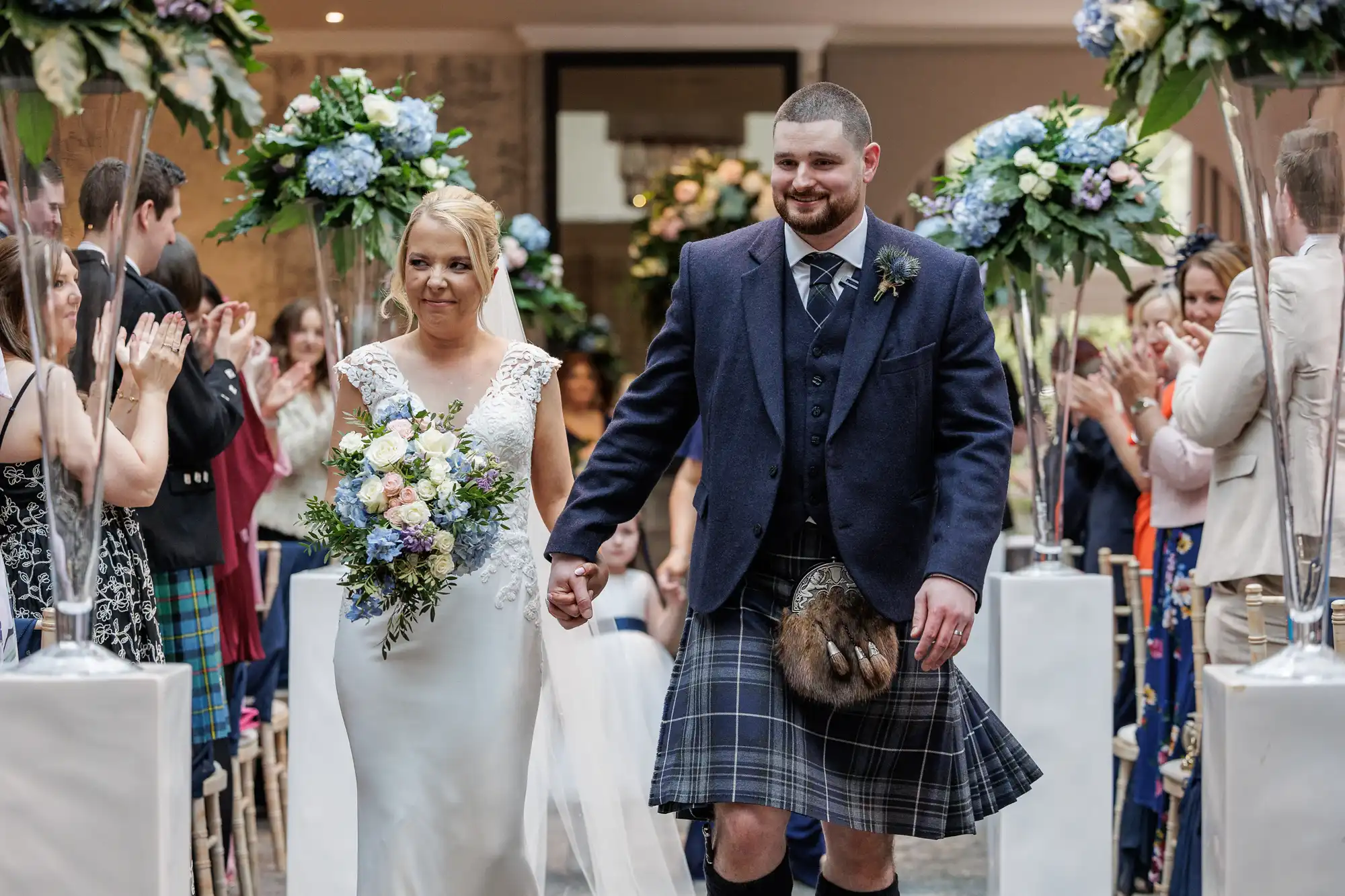 A bride in a white dress and a groom in a blue jacket and tartan kilt hold hands, walking down the aisle. Guests on both sides applaud, and tall vases with blue and white flowers line the aisle in a bright indoor venue.