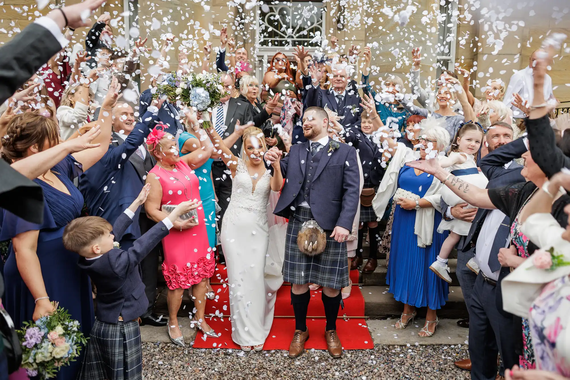 A bride and groom stand smiling on a red carpet surrounded by cheering guests, all showering them with white confetti. The groom wears a kilt; the bride wears a white dress. Friends and family in colourful outfits celebrate outdoors in front of a building.