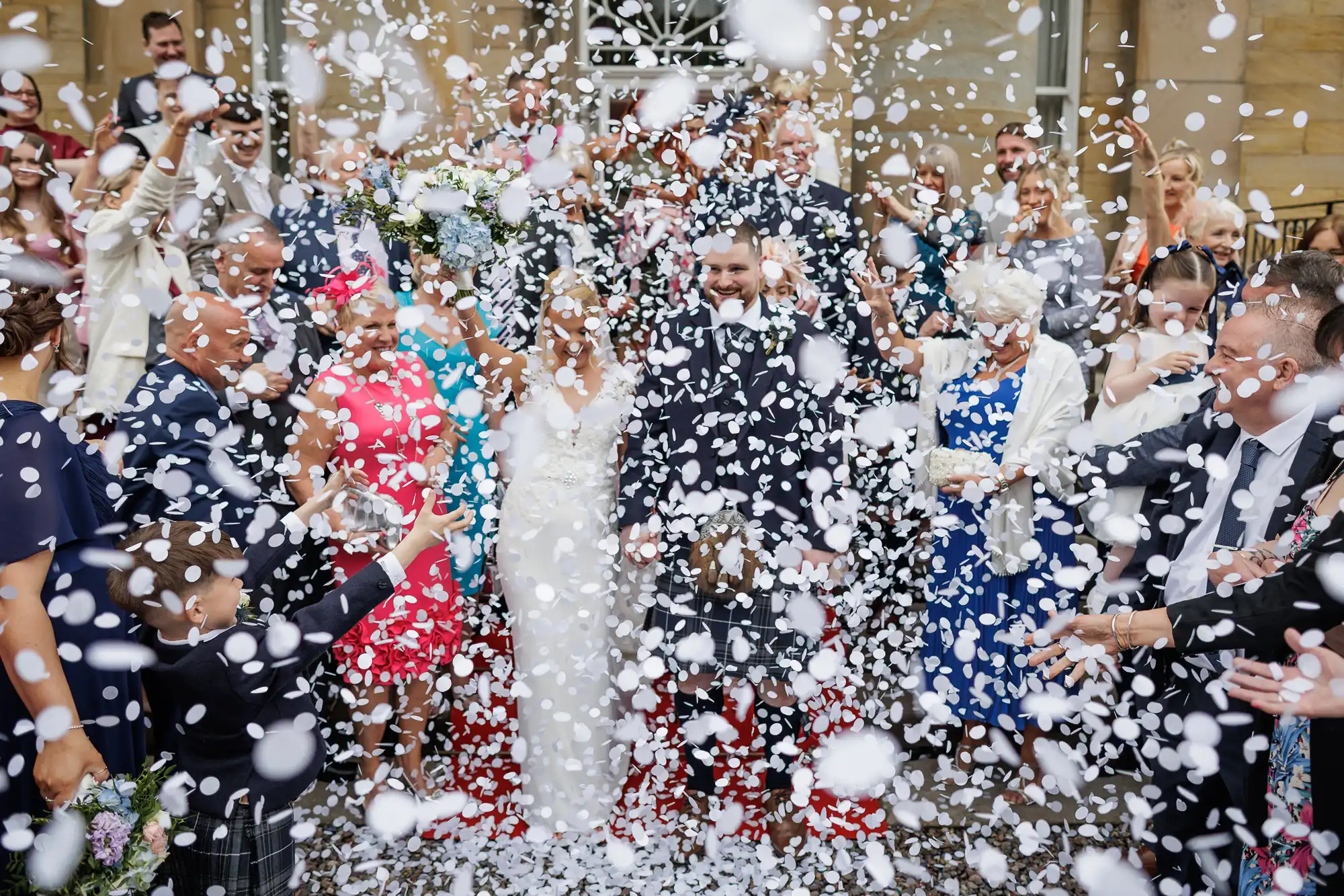 A bride and groom smile as they walk among guests throwing white confetti. The joyful crowd, dressed in formal attire, surrounds the couple outside a building. The confetti fills the air, partially obscuring faces but highlighting the celebration.