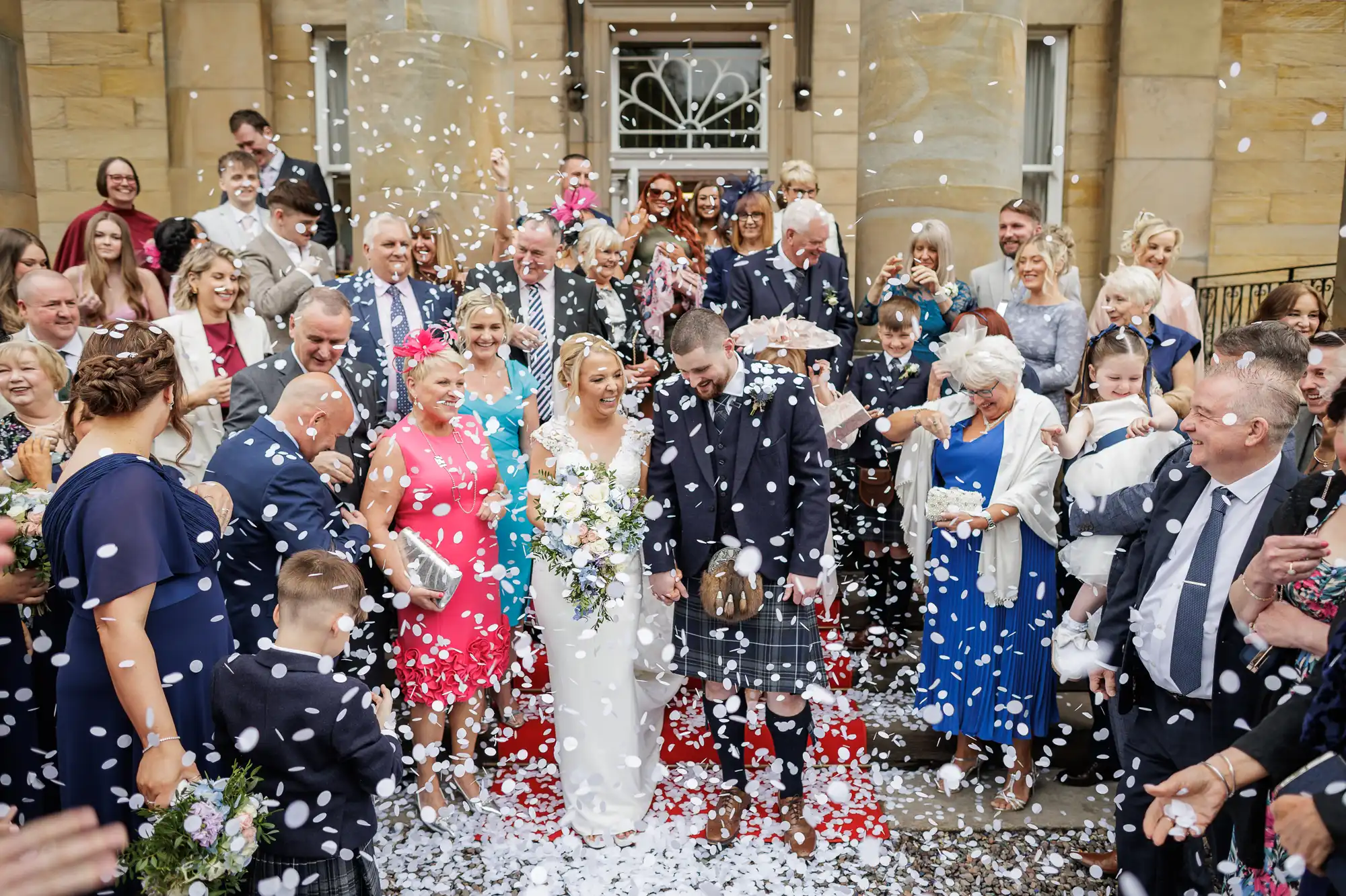 A bride and groom in wedding attire walk down a red carpet outside an elegant stone building, smiling as they are showered with white confetti. They are surrounded by cheering, well-dressed guests, some capturing the moment with mobiles and cameras.