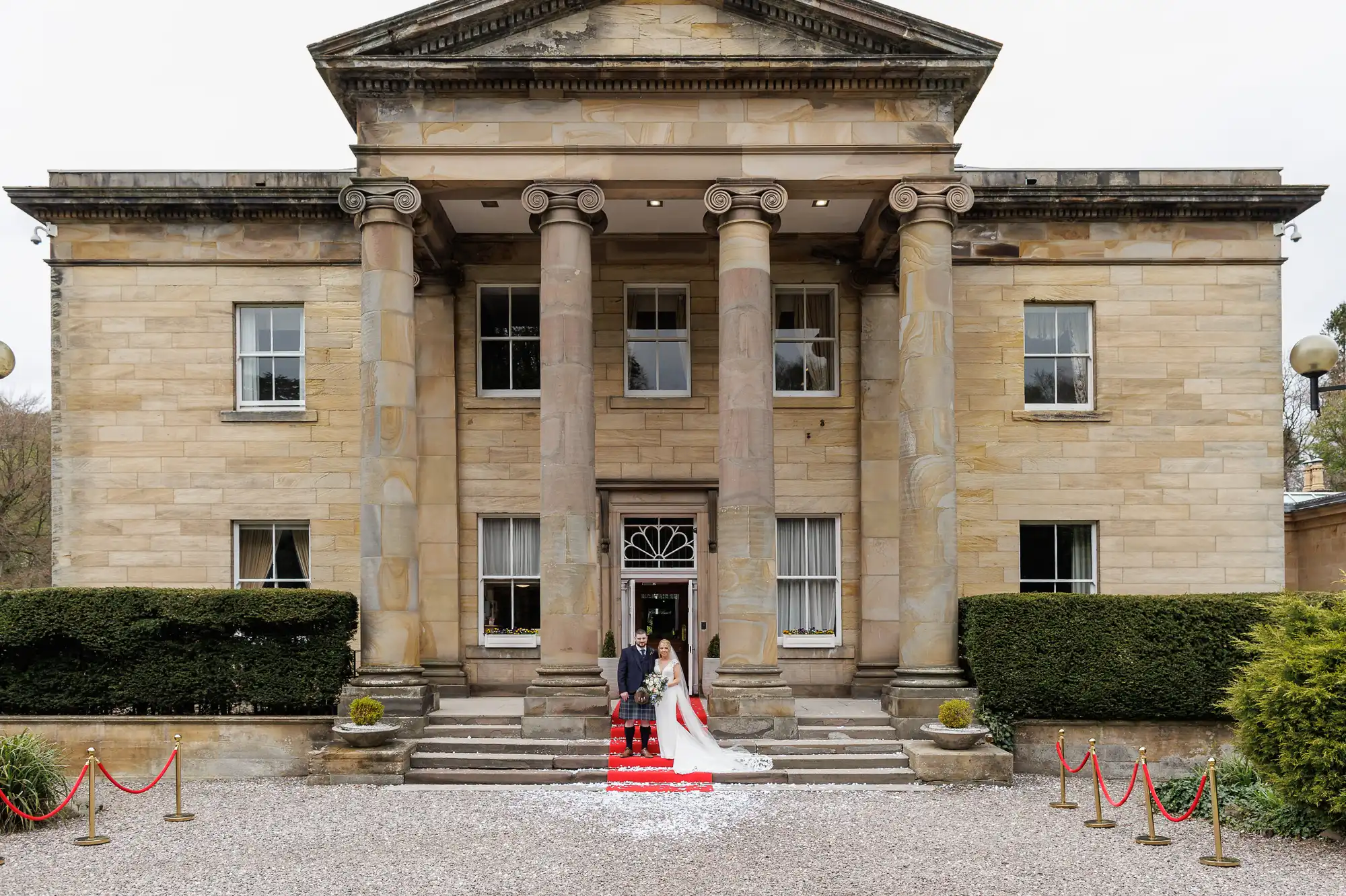 A bride in a white dress and a groom in a grey jacket with a red kilt stand on the front steps of a grand stone building with tall columns. Red rope barriers frame the gravel path leading to the entrance. Bushes flank the stairs.