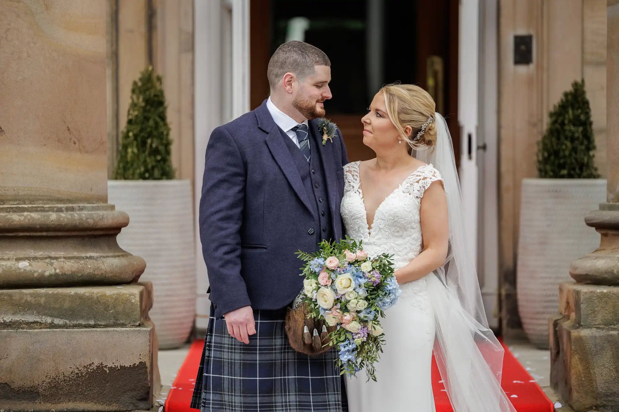 A bride in a white, lace wedding dress and veil holds a colourful bouquet, standing next to a groom in a dark jacket, waistcoat, tie, and tartan kilt. They gaze at each other, smiling, outside a building with stone columns and potted plants.