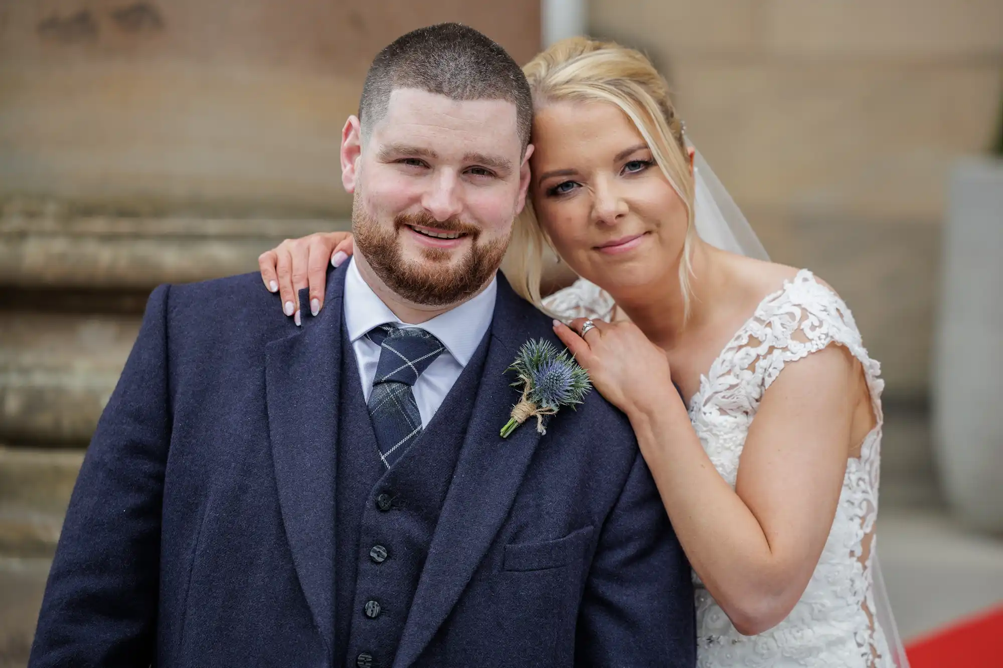 A smiling bride in a white lace gown and veil leans her head on the shoulder of a groom in a dark suit with a tartan tie and buttonhole. They stand close together outdoors, looking content and happy on their wedding day.