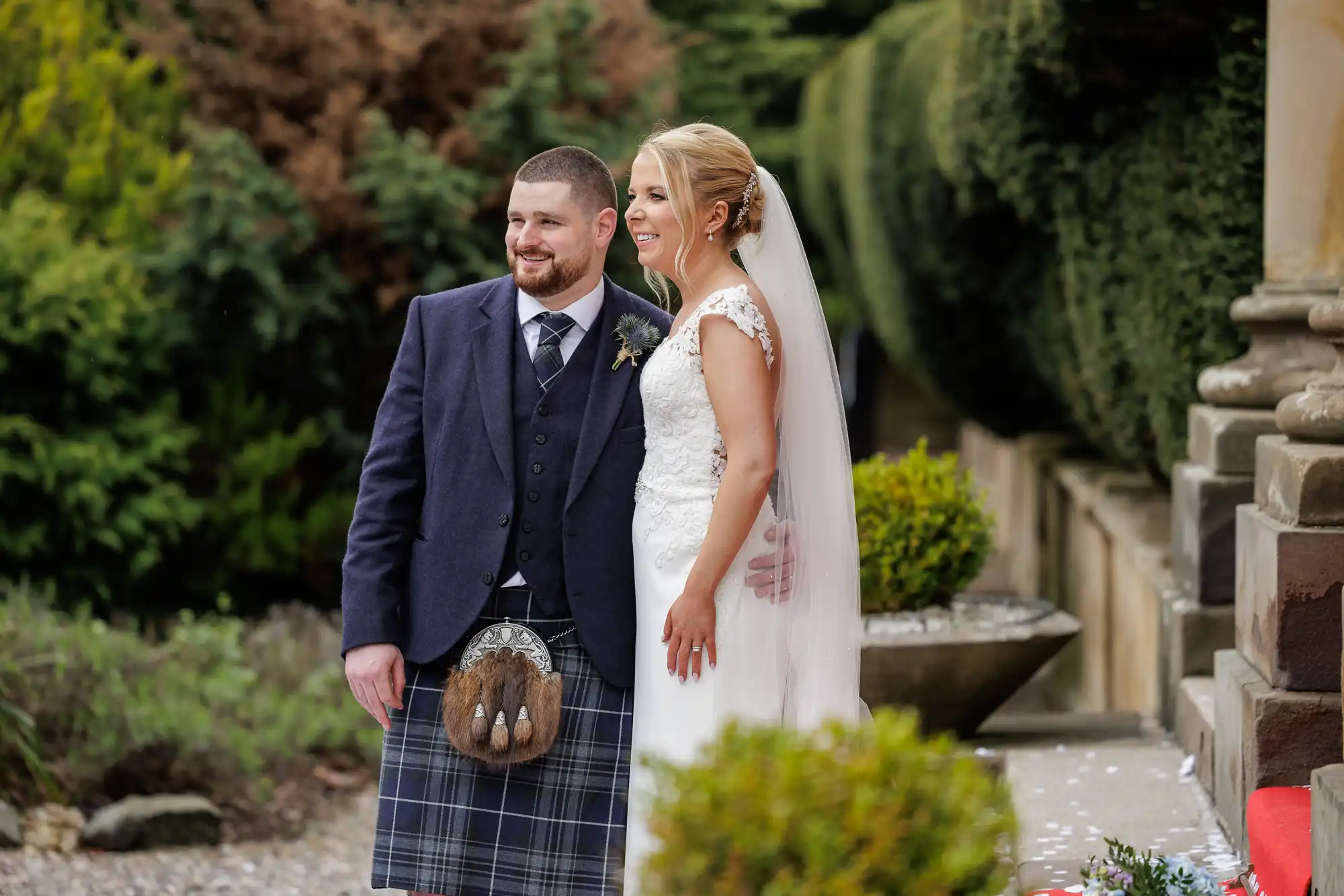 A smiling bride and groom pose outdoors. The groom wears a dark jacket, tartan kilt, and sporran; the bride wears a white dress with lace details and a long veil. They stand arm in arm near greenery and stone columns on a garden path.