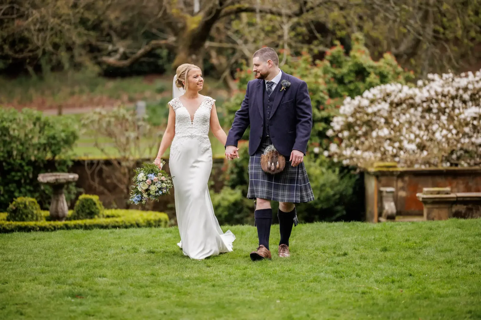A bride in a white gown and a groom in a navy jacket and tartan kilt walk hand-in-hand on green grass in a garden, surrounded by flowering bushes and trees, gazing happily at each other. The bride holds a bunch of flowers.