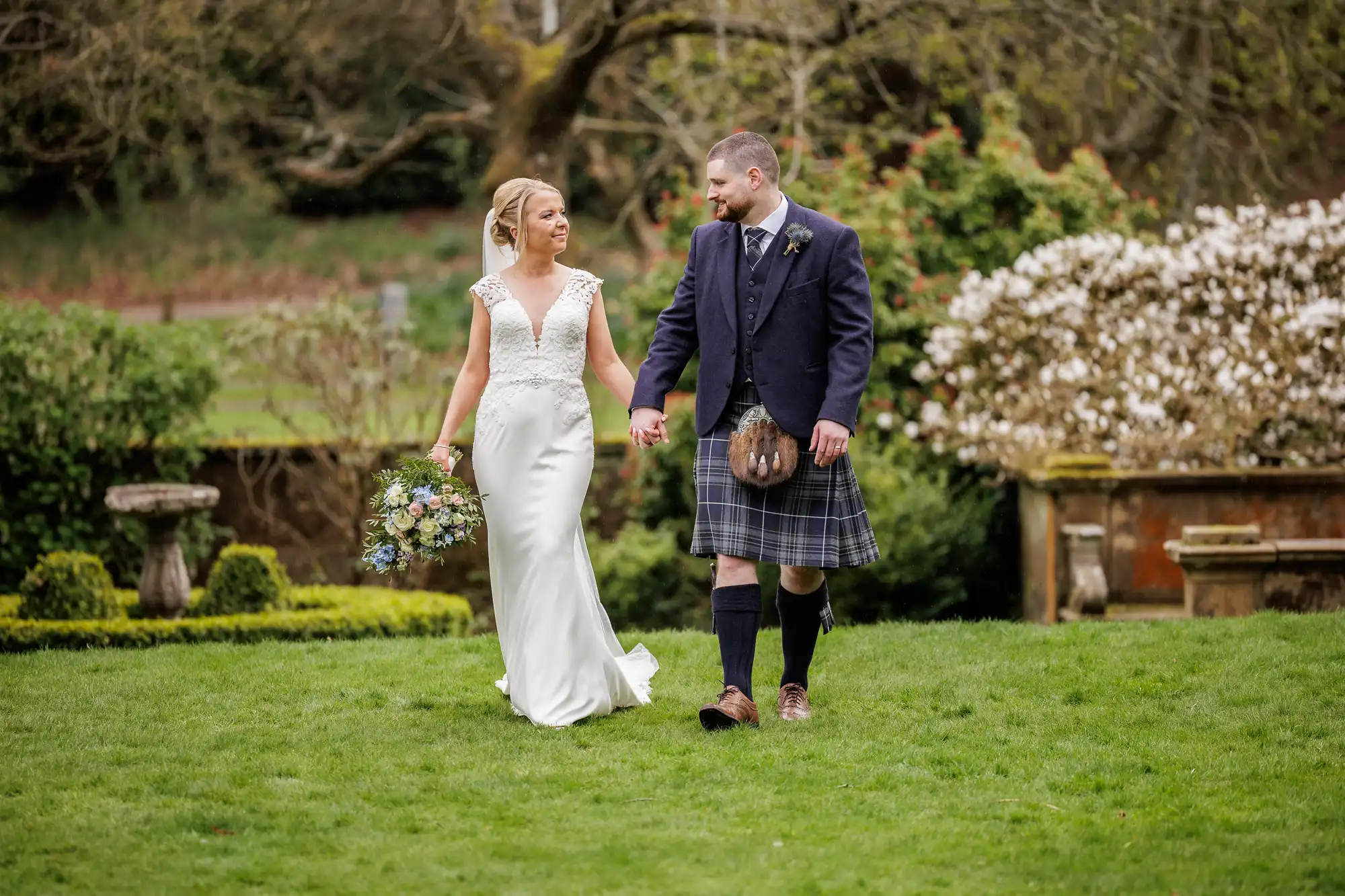A bride in a white gown and a groom in a navy jacket and tartan kilt walk hand-in-hand on green grass in a garden, surrounded by flowering bushes and trees, gazing happily at each other. The bride holds a bunch of flowers.