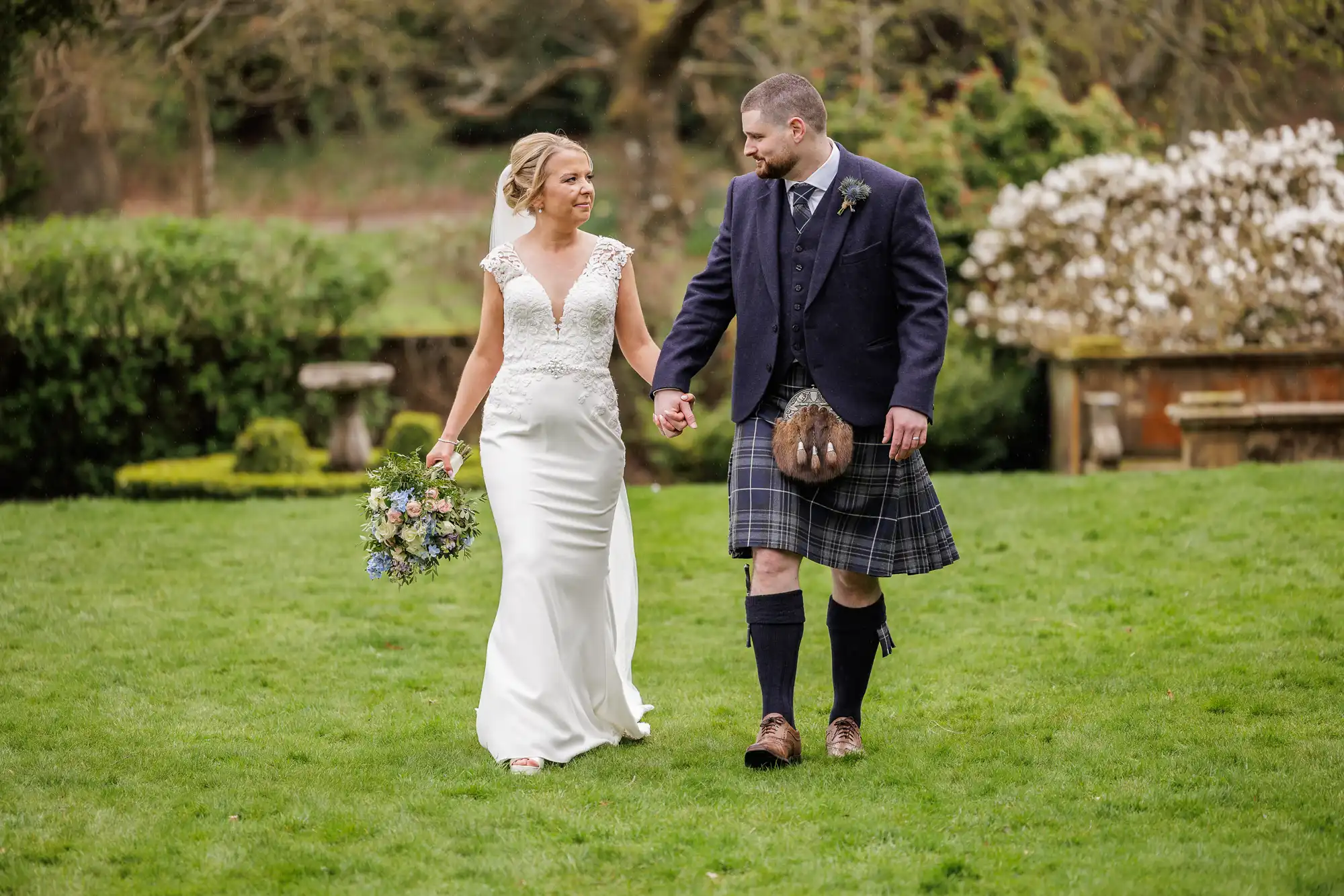 A bride in a white dress holding a bouquet and a groom in a dark jacket, waistcoat, and traditional tartan kilt walk hand in hand on green grass in a garden, smiling at each other. Blossoming bushes and trees are visible in the background.