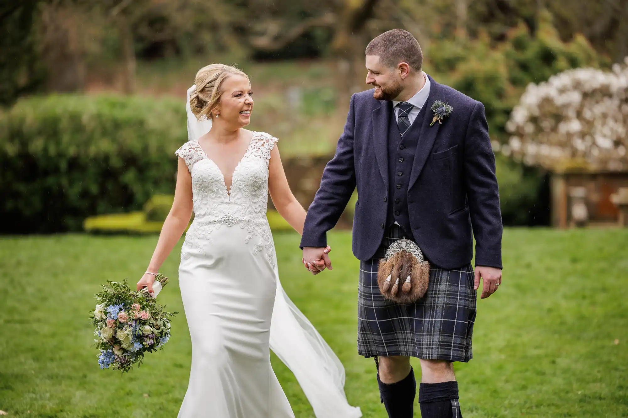 A bride in a white gown holding a bouquet and a groom in a navy jacket and tartan kilt smile at each other whilst holding hands, walking on green grass outdoors, with blurred trees and bushes in the background.