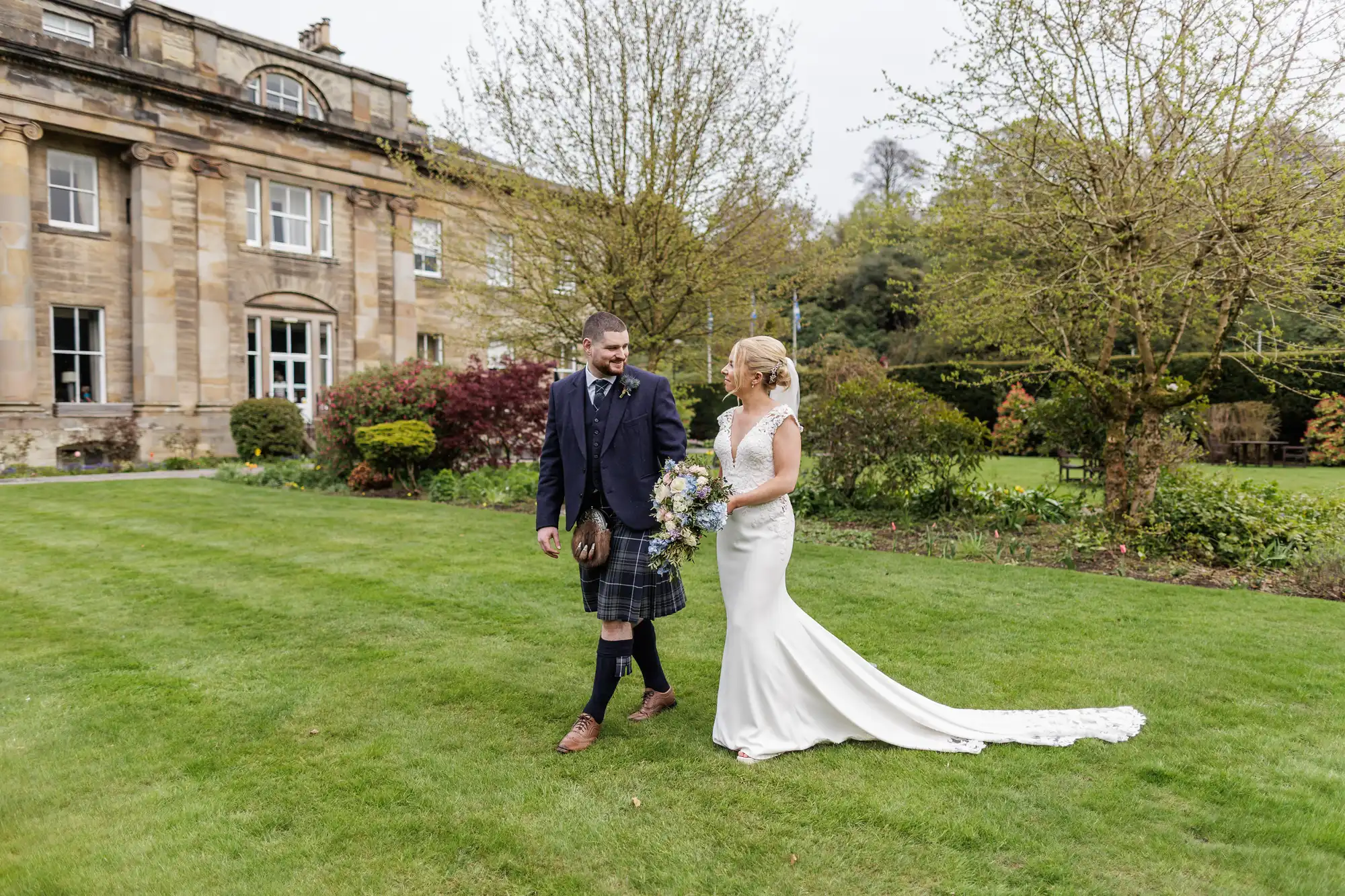 A bride in a white dress and a groom in a kilt walk hand in hand on a green lawn, smiling at each other. Behind them is a grand stone building and colourful landscaped gardens with trees and shrubs.
