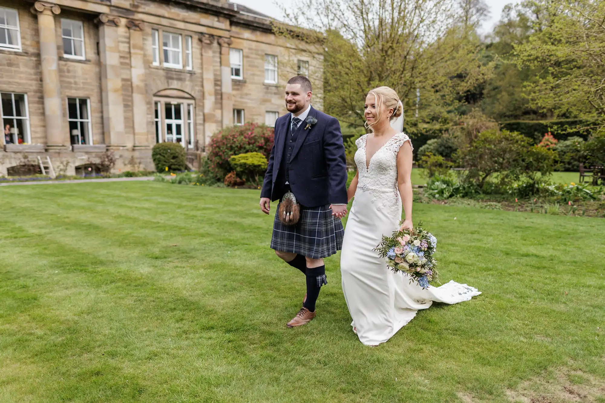 A smiling couple walk on grass outside a large stone building. The woman wears a white wedding dress and holds a bouquet; the man wears a blue jacket and tartan kilt. Trees, bushes, and spring flowers surround them in a garden setting.