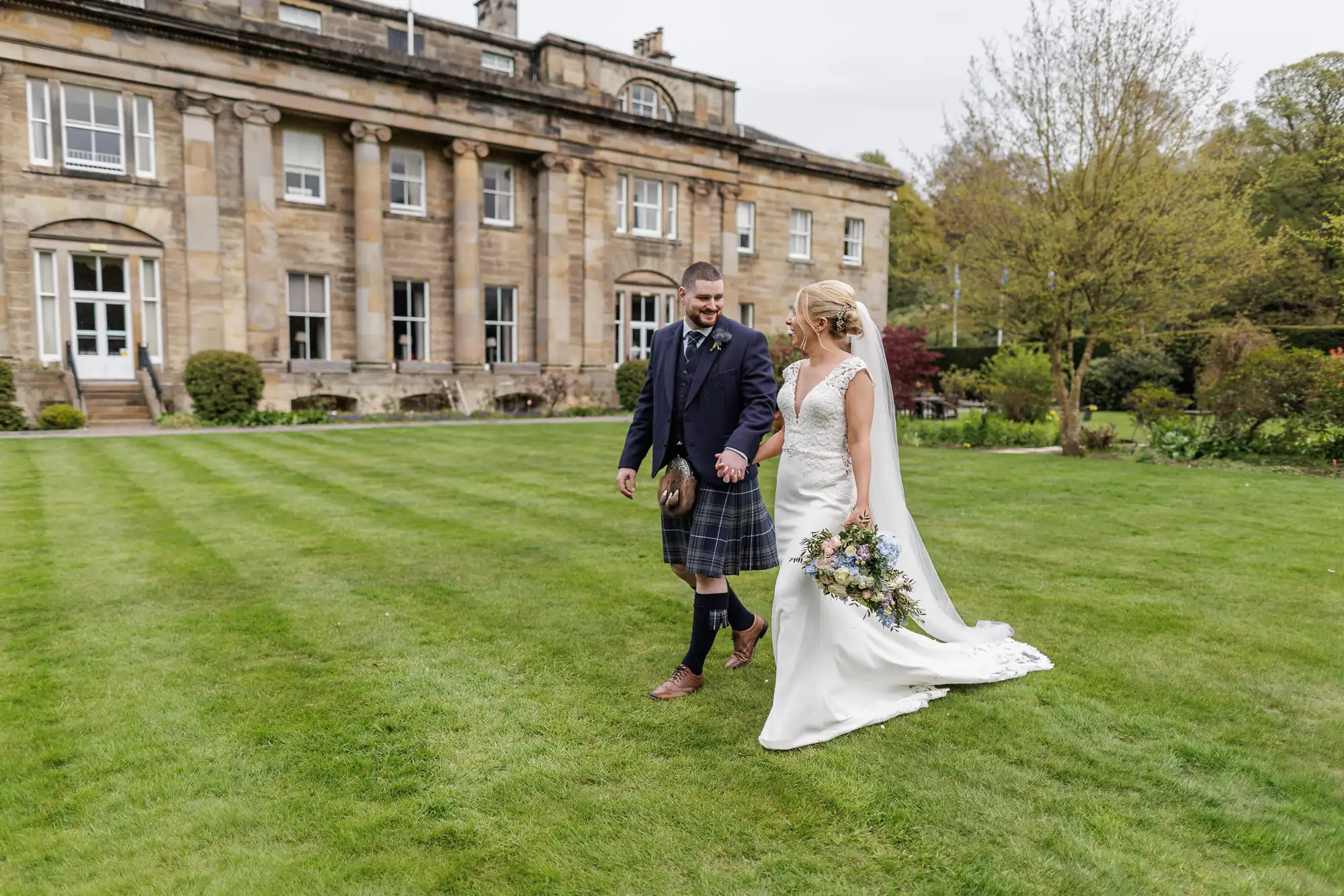 A bride in a white gown holding a bouquet walks hand-in-hand with a groom in a dark jacket and kilt. They are smiling at each other in front of a large stone manor, surrounded by green, manicured lawns and trees.