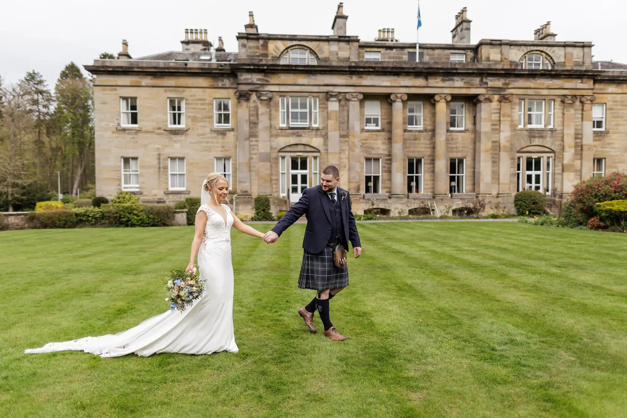A bride in a white gown and a groom in a kilt walk hand in hand across a green lawn in front of a grand stone manor, surrounded by manicured shrubs. The bride holds a bouquet and both are smiling, enjoying their wedding day outdoors.
