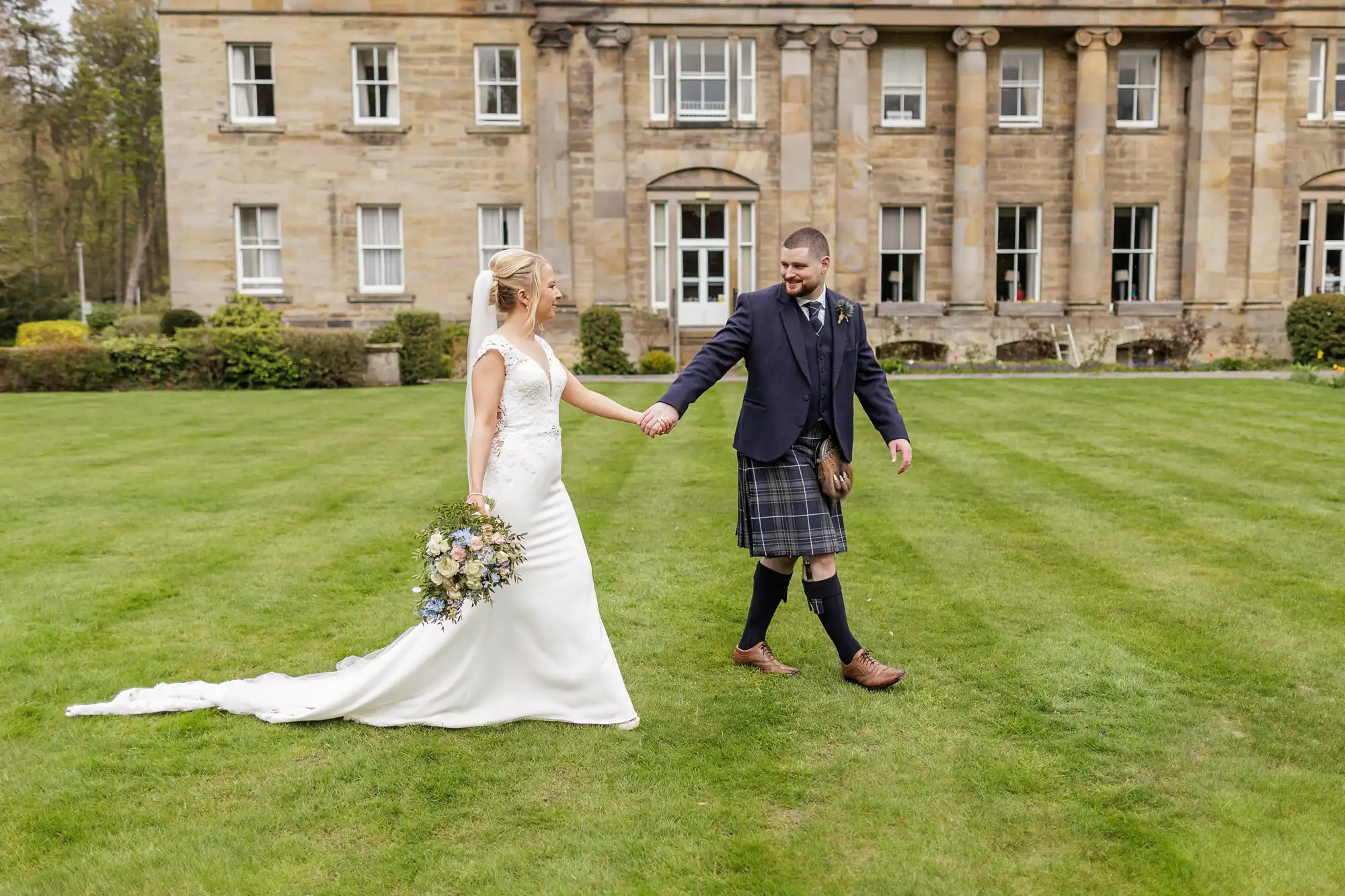 A bride in a white dress holds a bouquet and walks hand-in-hand with a groom in a dark jacket and kilt, across green grass in front of a grand stone building with tall windows and columns.