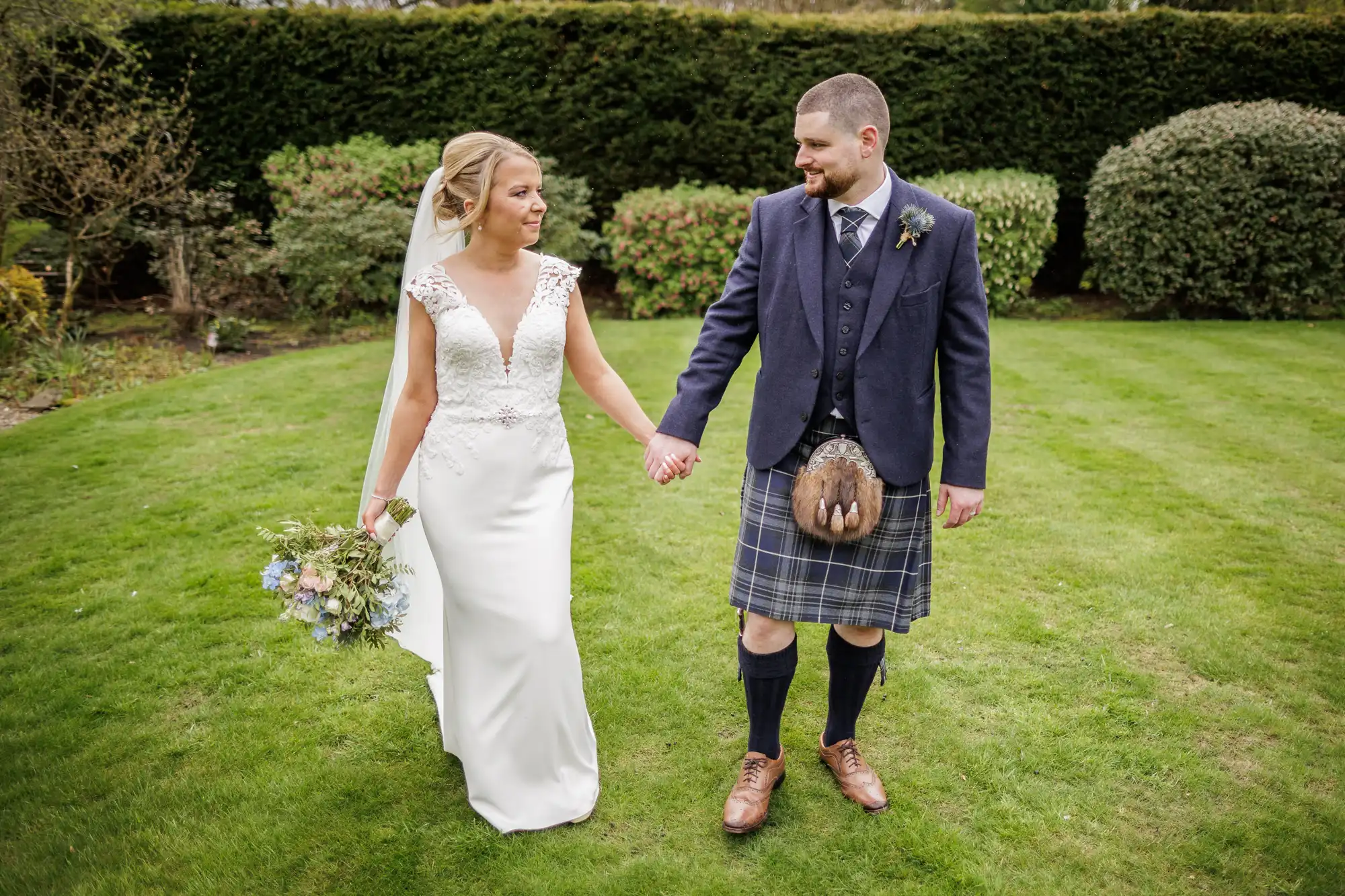 A bride in a white dress and veil holds a bouquet and the hand of a groom in a dark jacket and traditional Scottish kilt. They smile at each other whilst walking on green grass, with bushes and trees in the background.