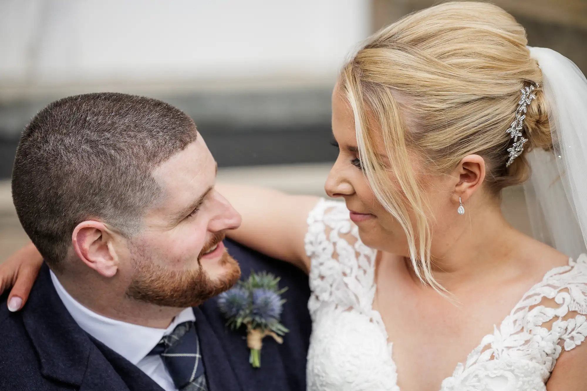 A bride and groom gaze lovingly at each other. The groom has short brown hair, a beard, and wears a dark suit with a buttonhole. The bride, with blonde hair in an updo and a veil, wears a white lace dress and pearl earrings. They both smile softly.