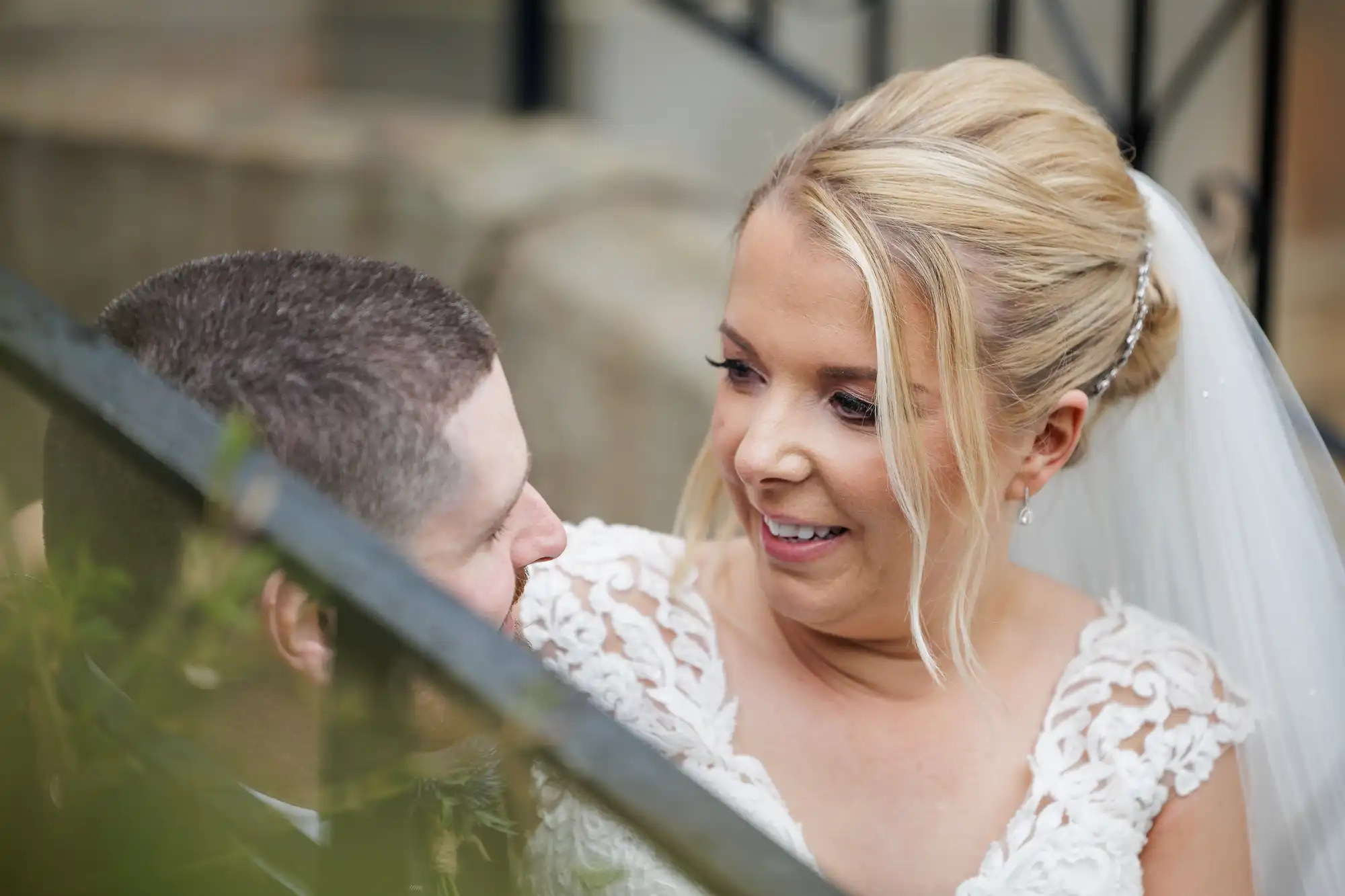 A blonde bride in a white lace dress and veil smiles warmly at a groom with short brown hair and a beard. They are closely seated outdoors, gazing at each other lovingly, with blurred greenery and metal railings in the foreground.