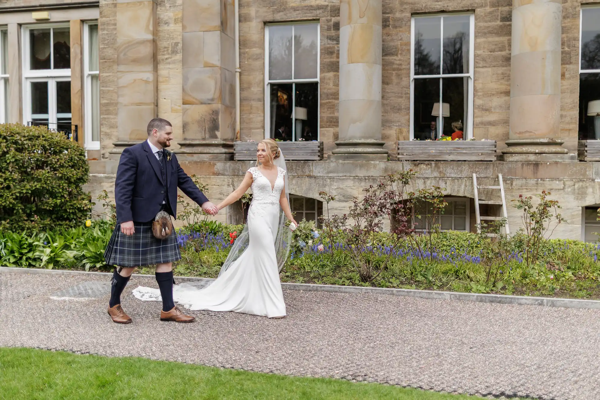 A bride in a white dress and a groom in a dark jacket, tie, and a checked kilt walk hand in hand outside a historic stone building with large windows, columns, and a manicured garden. Both are smiling and looking at each other.
